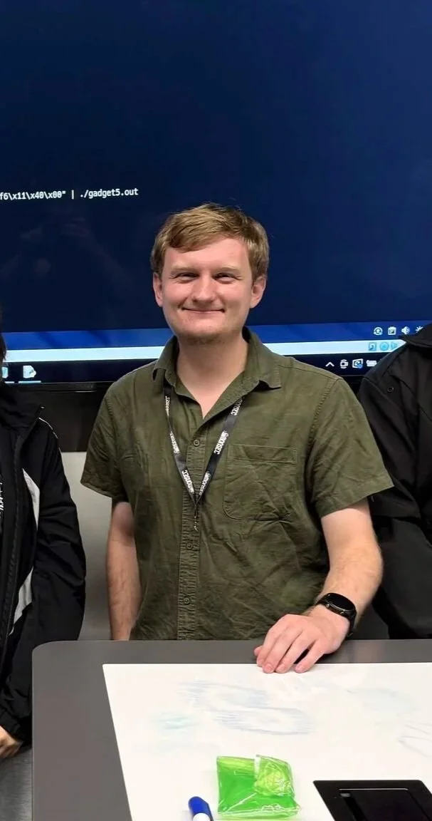 A smiling young man with light brown hair, wearing a green short-sleeved button-up shirt, standing behind a table with a white paper and a green plastic bag. He is in front of a large computer monitor.