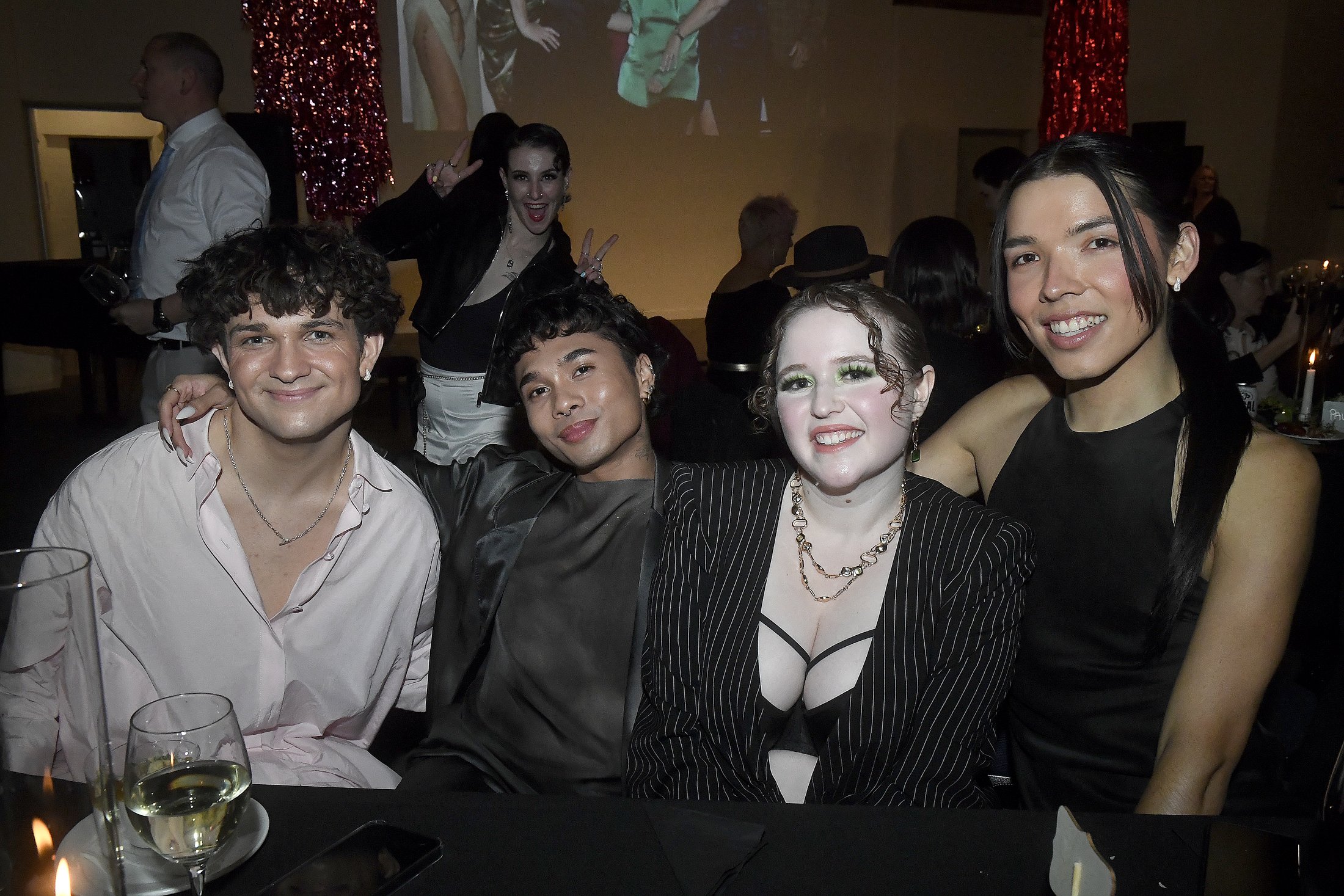 Group of four diverse friends smiling and posing together at a party, with a woman making a peace sign in the background.
