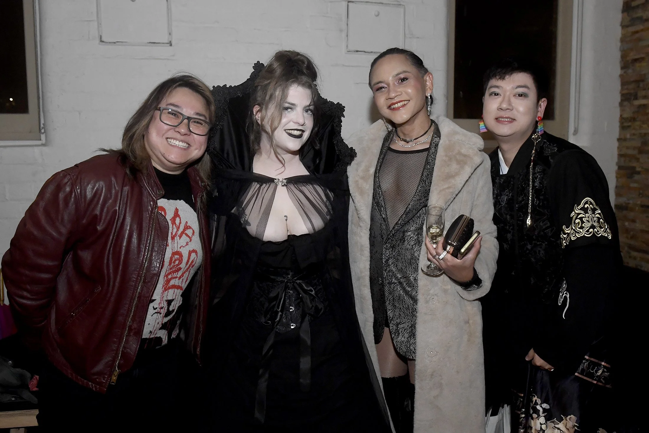 A group of five diverse women posing together indoors at a social gathering, smiling at the camera, with some holding drinks.