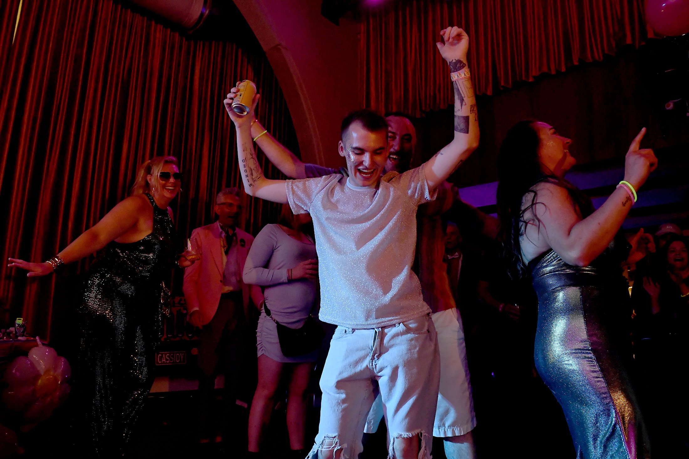 Young man dancing and smiling with arms raised among a group of people at a party or celebration in a dimly lit room with red curtains.