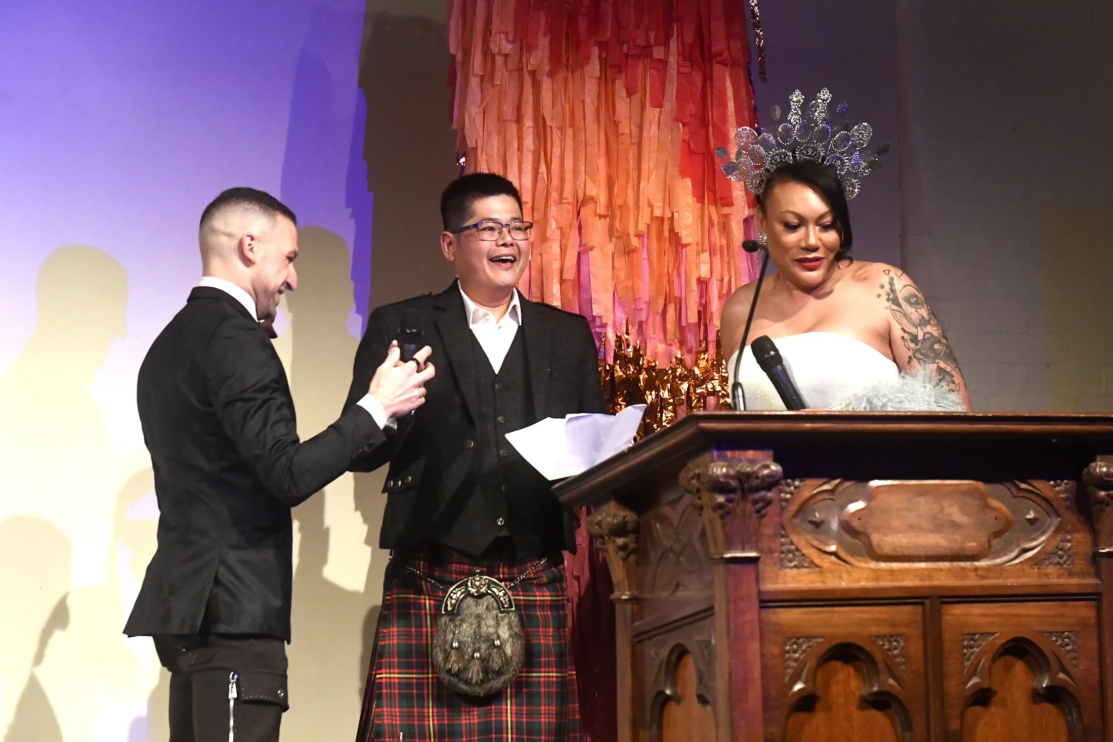 A woman wearing a crown and white dress stands at a wooden podium with a microphone, while two men in suits and a kilt laugh nearby during a celebration or ceremony.