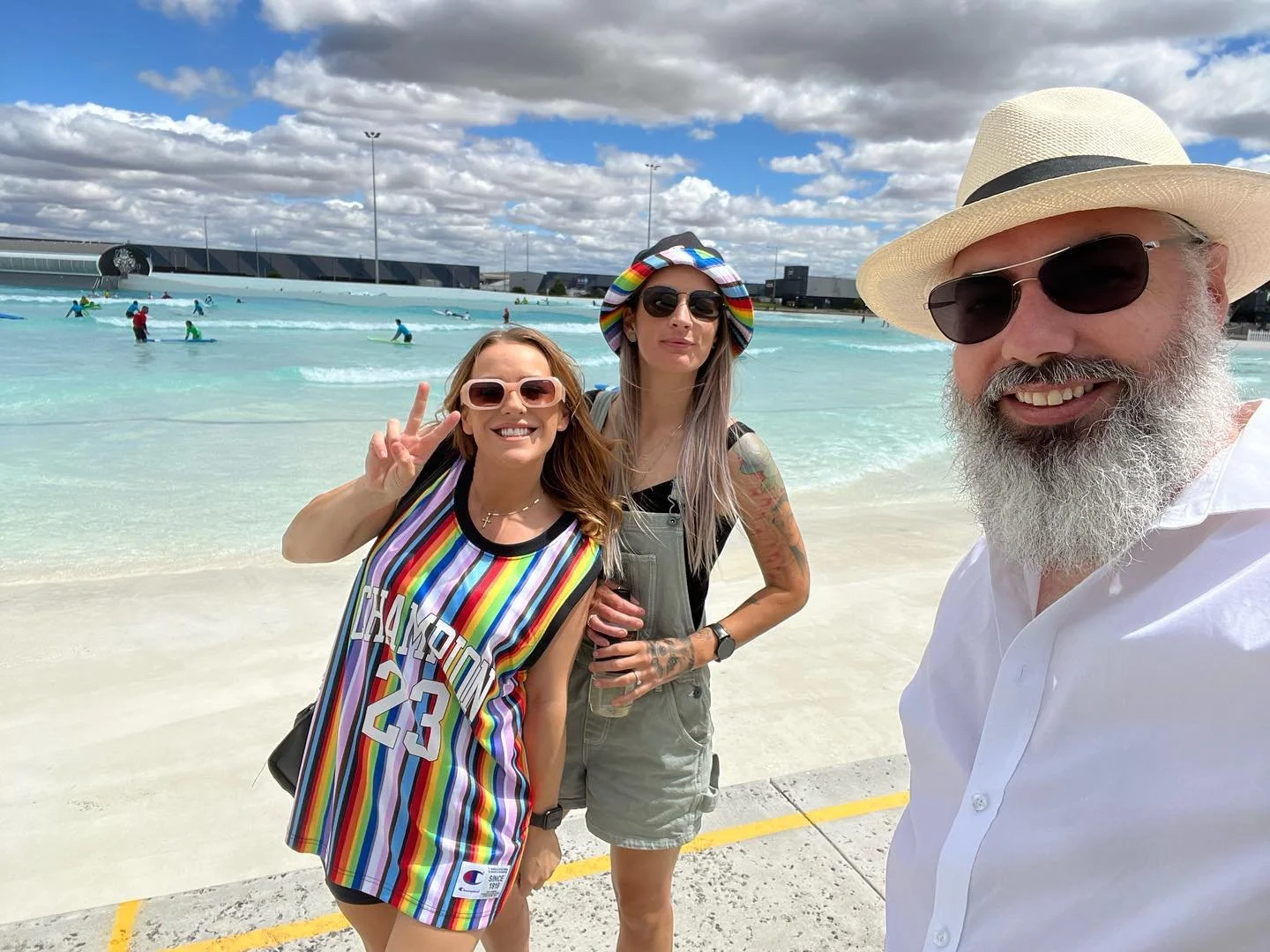 Three people taking a selfie on a beach with a wave pool, some people swimming, and a cloudy sky in the background.