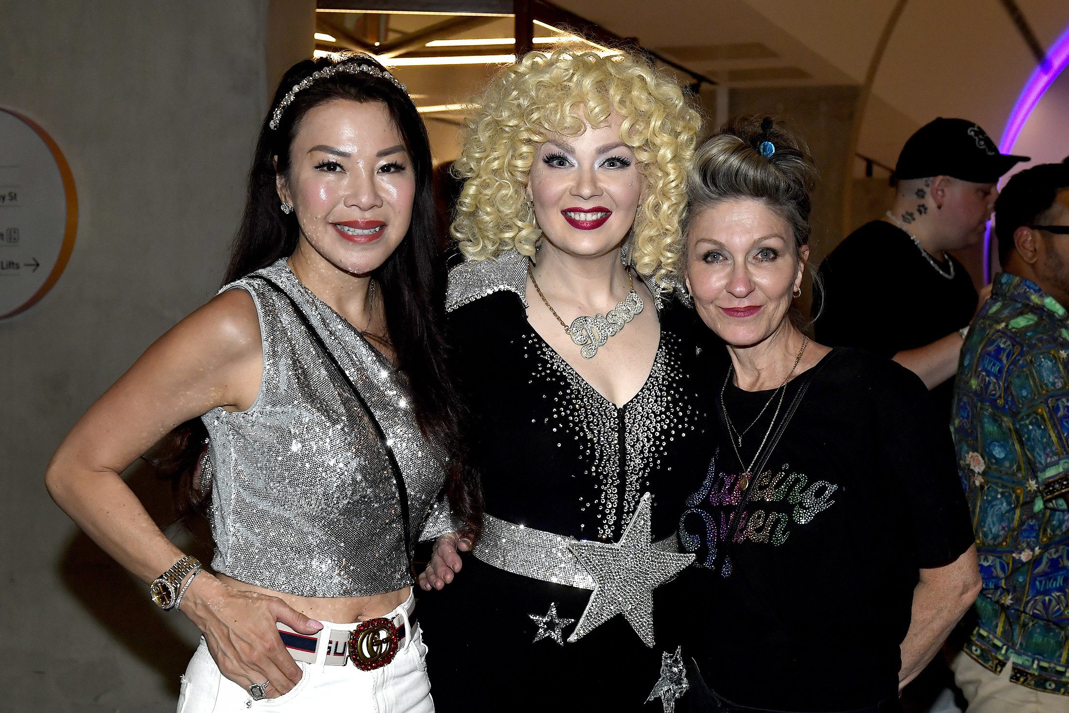 Three women dressed in shiny, glamorous clothing posing together at an indoor event.