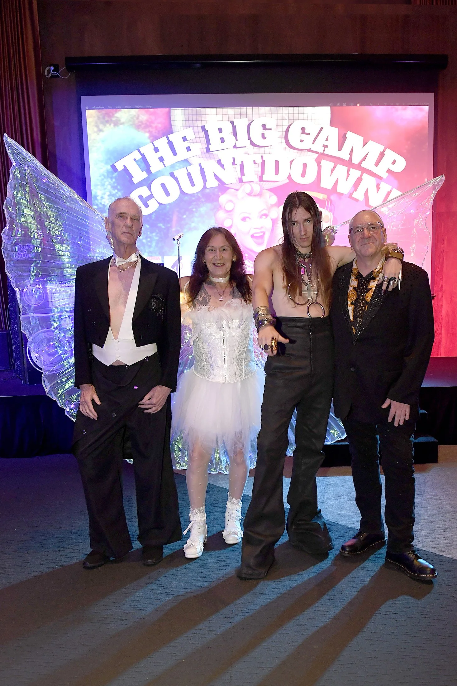 Four individuals dressed in eclectic costumes standing in front of a sign that reads 'The Big Camp Countdown' with angel wings and a cartoonish face in the background.