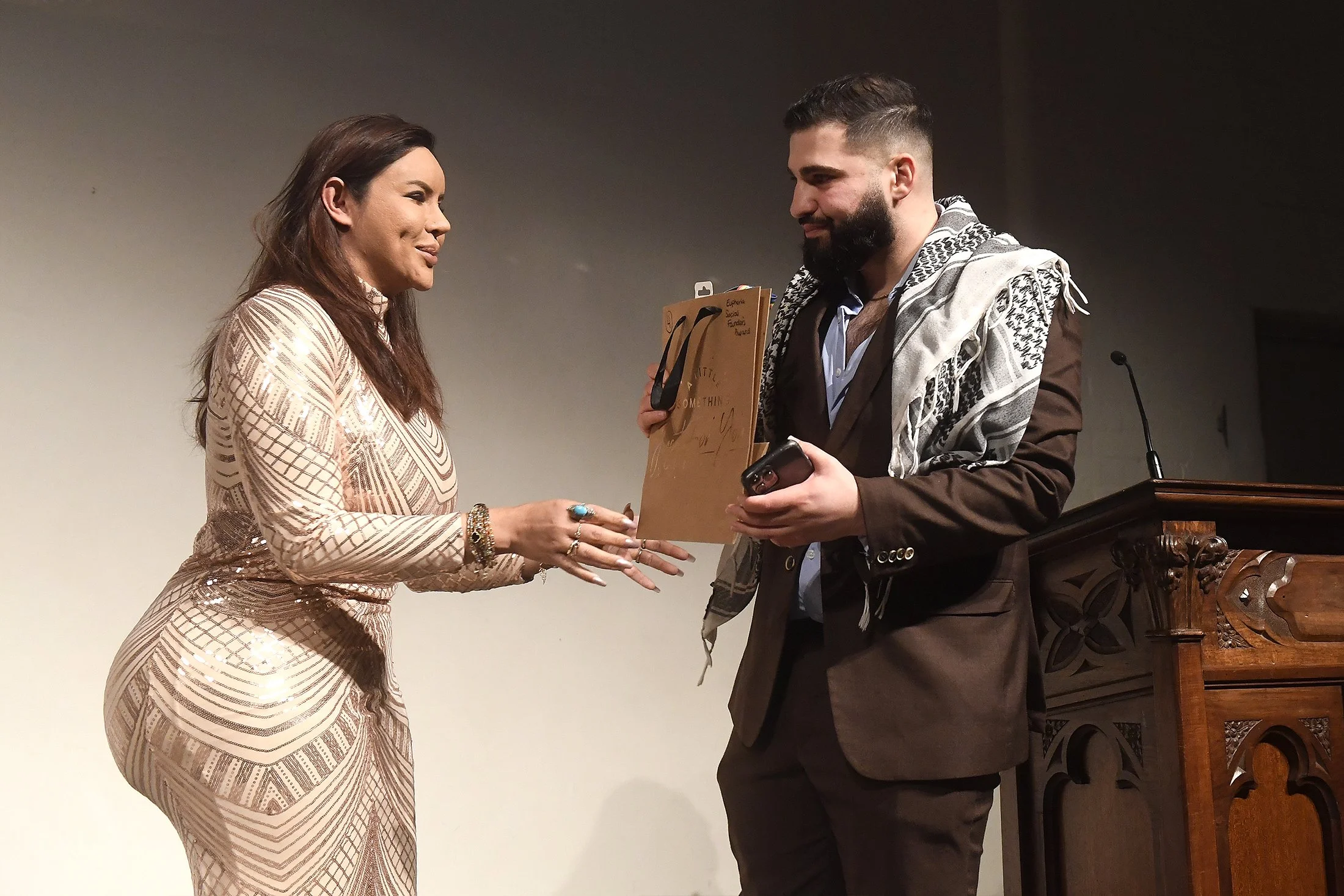 A woman in a shiny, patterned dress is receiving an award or certificate from a man in a brown suit with a patterned scarf. They are standing in front of a plain wall with a podium nearby.