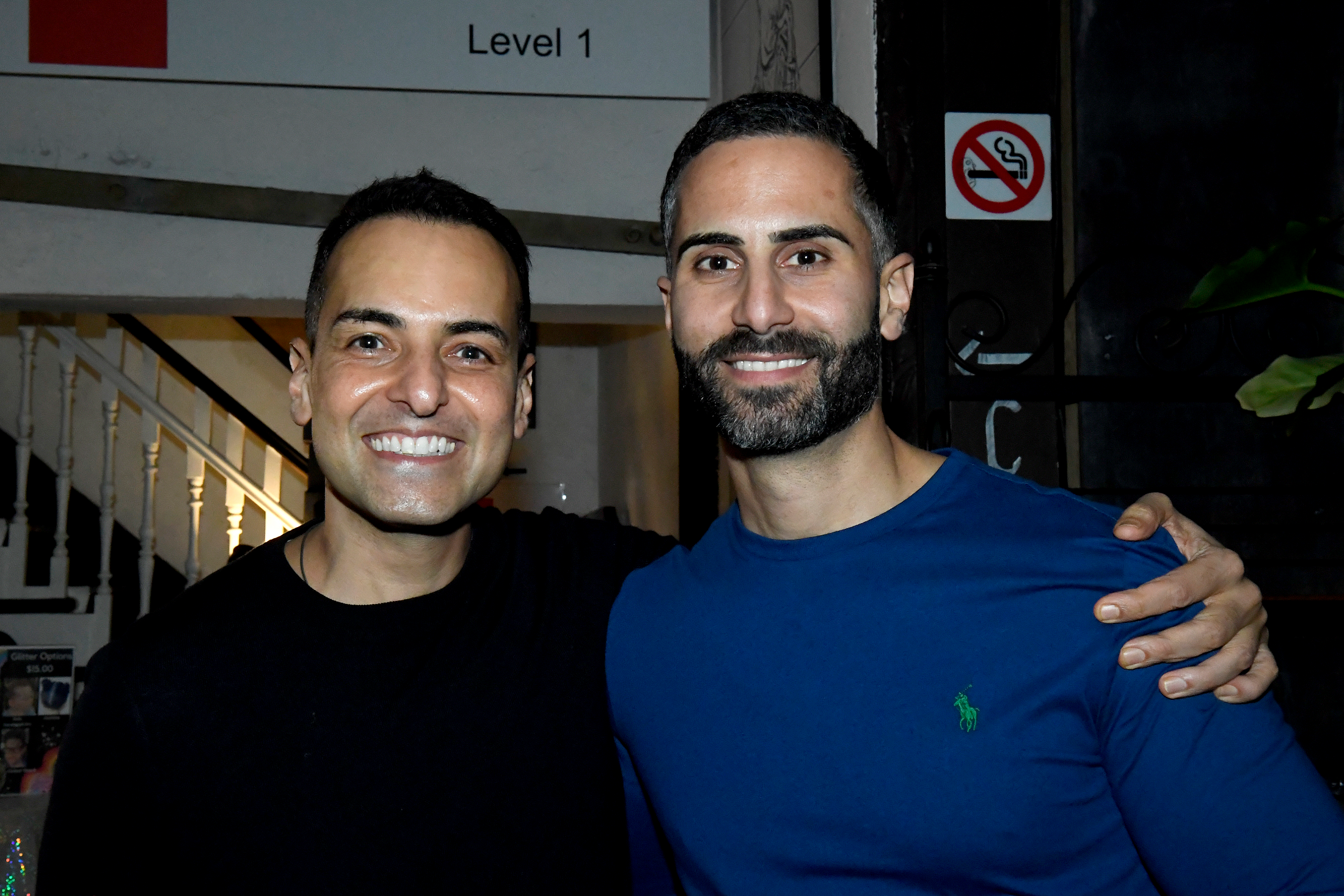 Two smiling men with dark hair and beards embracing each other indoors, one wearing a black shirt and the other a blue shirt, with a staircase and no smoking sign in the background.