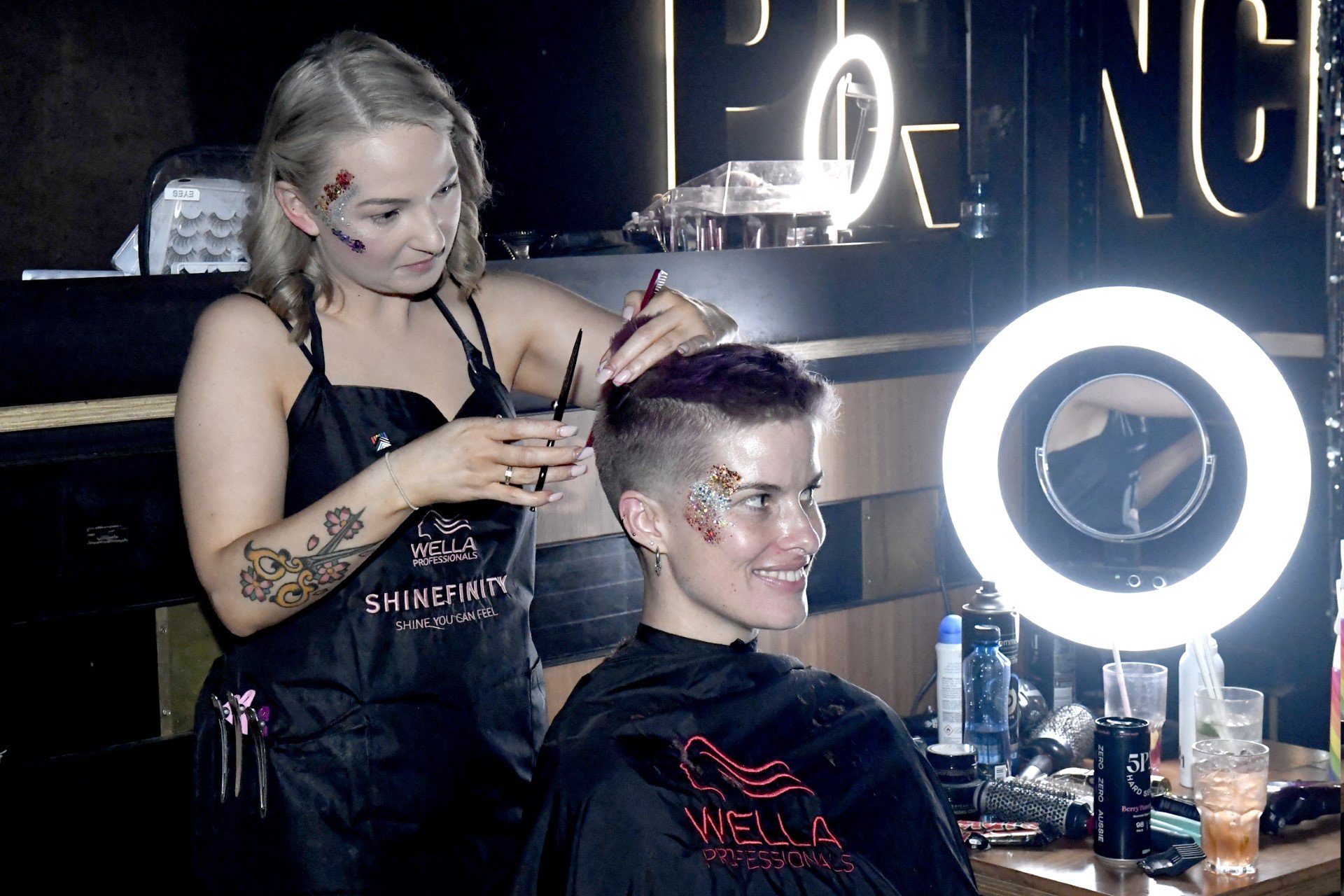 A hairstylist with tattoos and glitter face art cuts a woman's hair in a salon. The woman, also with glitter face art, smiles as she gets a fresh haircut. There are beauty products and a ring light on the table near them.