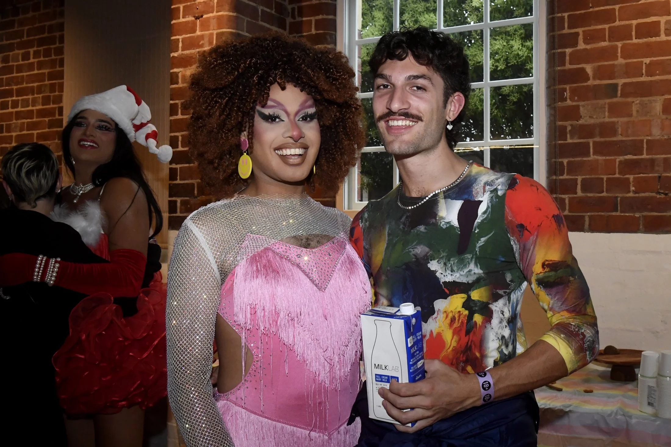 Two people smiling at a party, with a woman dressed in a colorful outfit and a man holding a milk carton, festive decorations, and another person in the background wearing a Santa hat.