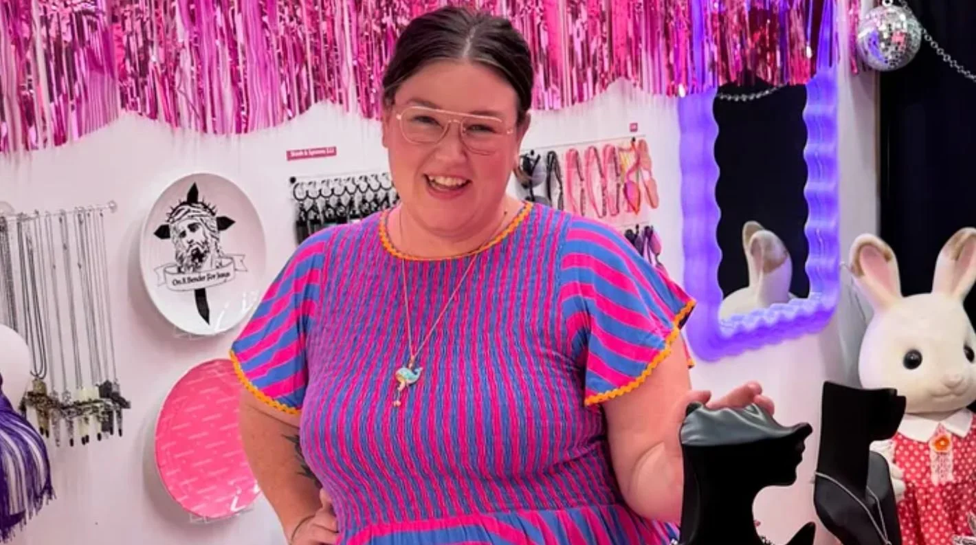 Woman with glasses and a pink and blue striped dress smiling in a store filled with accessories and decorations, including bunny head mannequins and colorful hanging streamers.