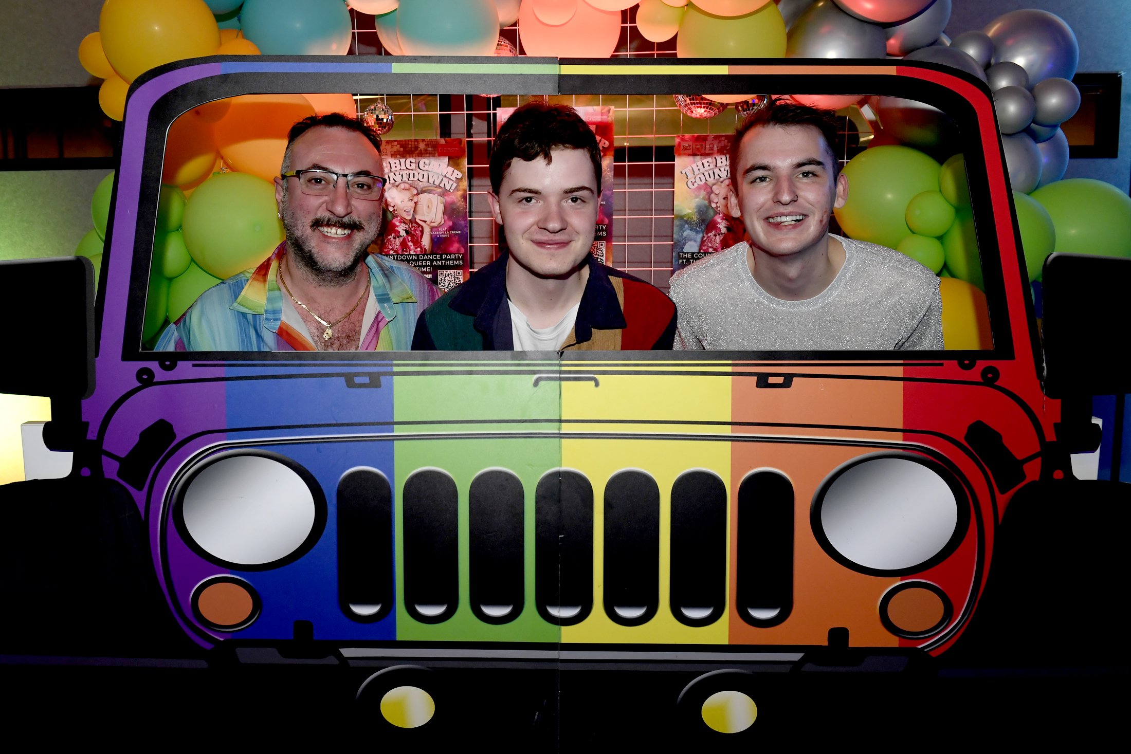 Three people are sitting behind a cutout of a rainbow-colored Jeep with a rainbow background of balloons and posters.