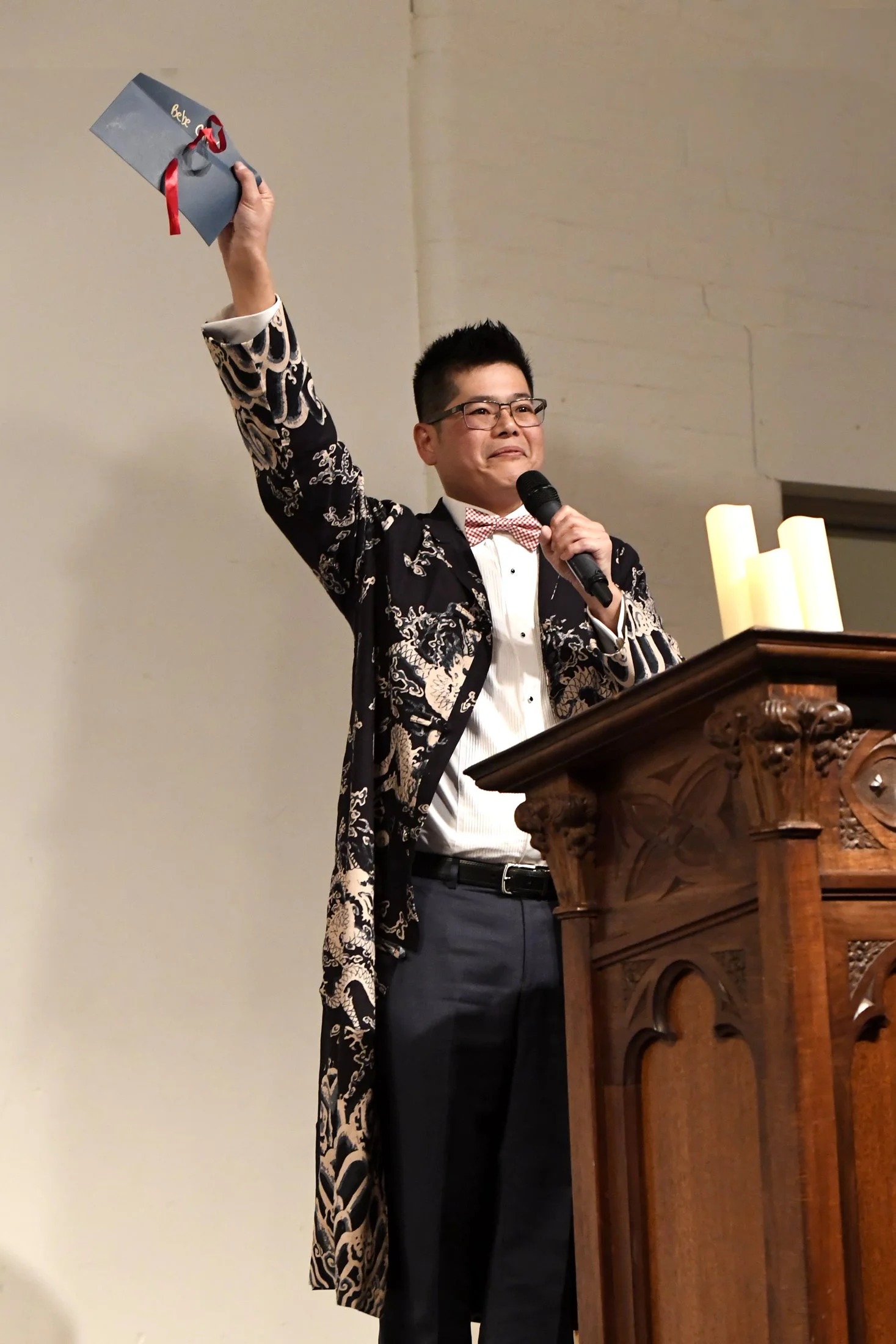 A man in formal attire, including a patterned robe, white shirt, and red checkered bowtie, stands at a wooden pulpit, holding a microphone in one hand and a gift box with a red ribbon in the other, in a church or similar setting.
