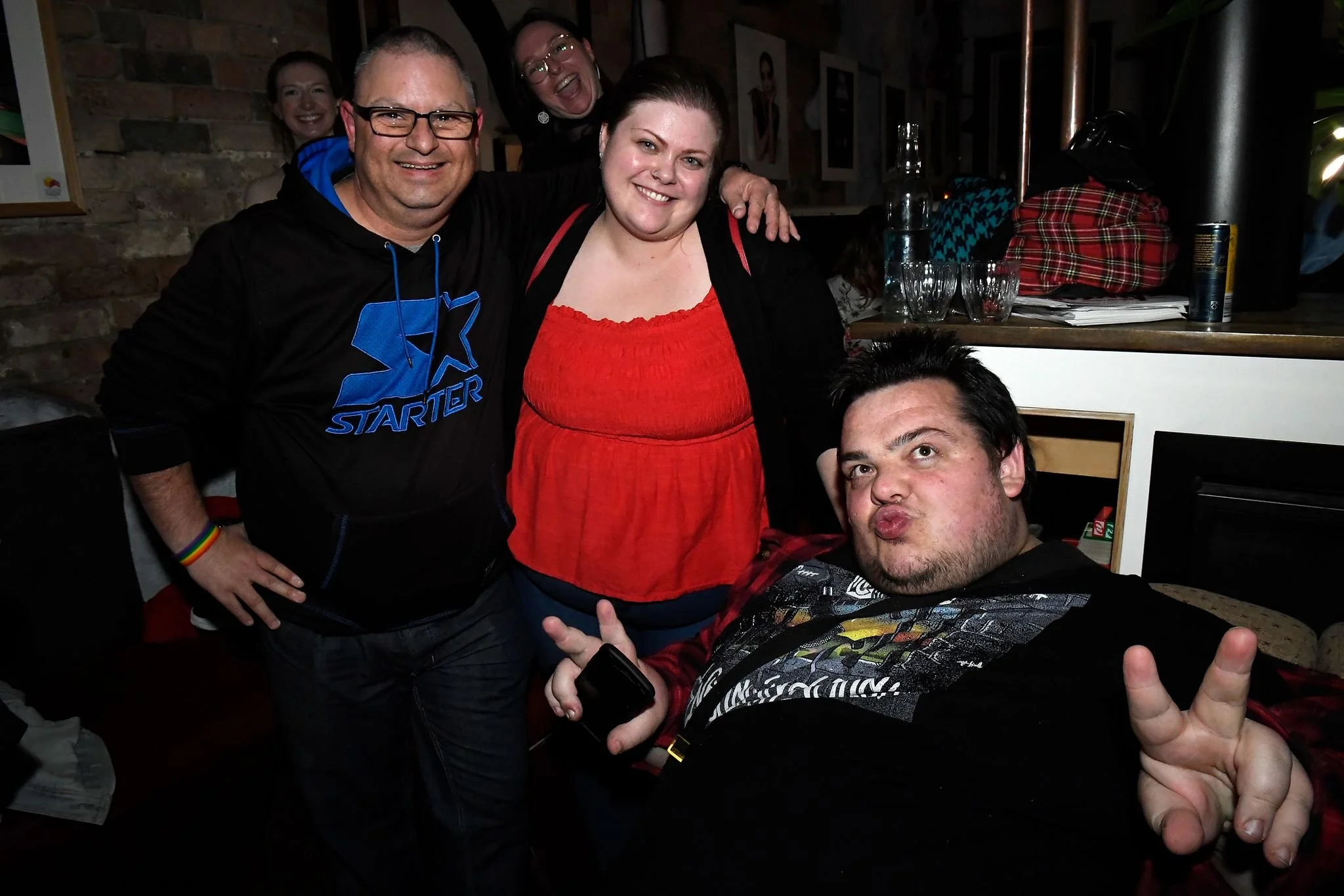 Group of four friends smiling and making peace signs at a social gathering in a cozy indoor setting.