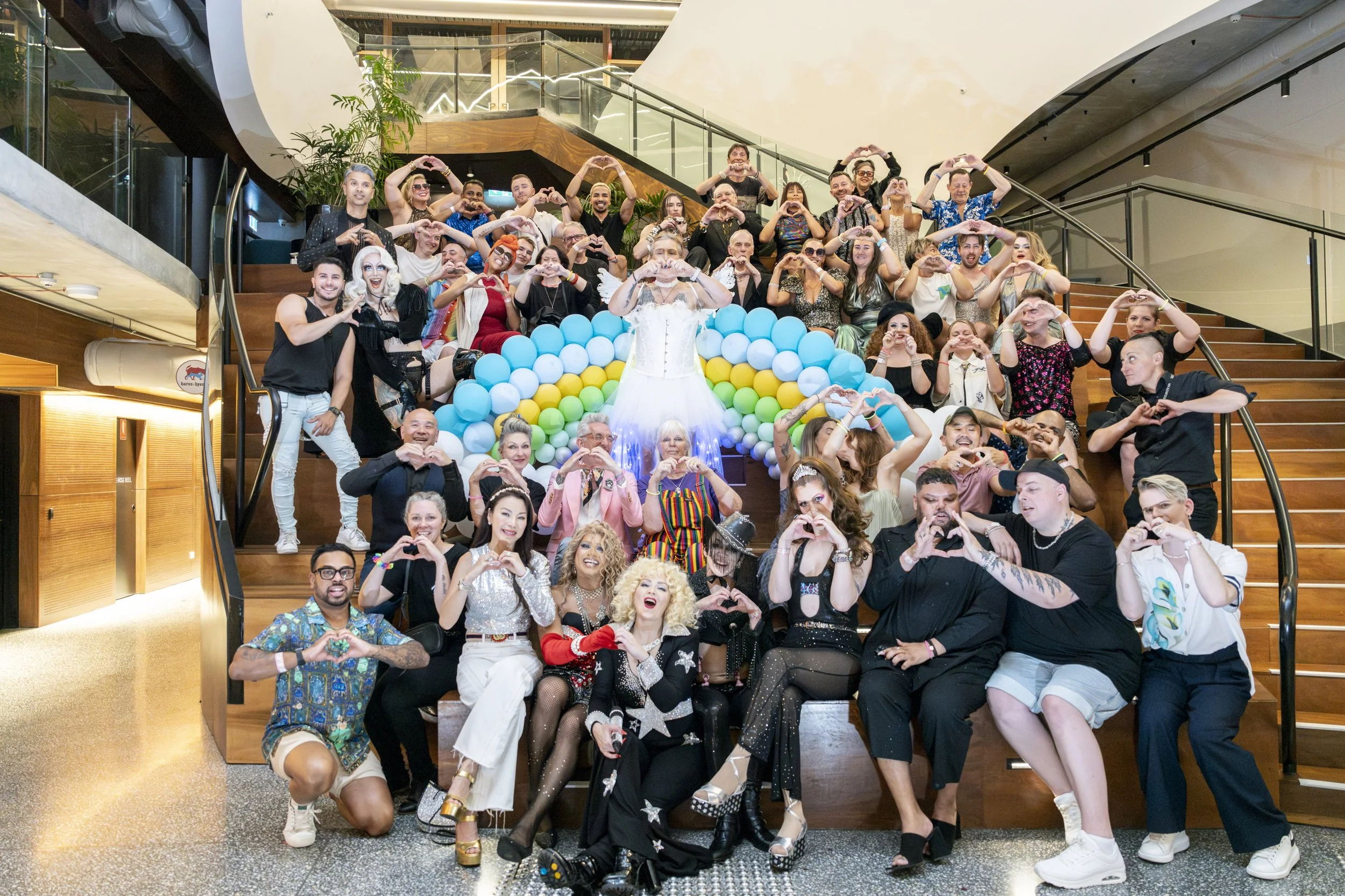A large group of people gathered on and around a staircase, holding heart shapes with their hands. The background features a large rainbow-shaped balloon display and a white dress worn by a person at the center. The group appears to be celebrating, w