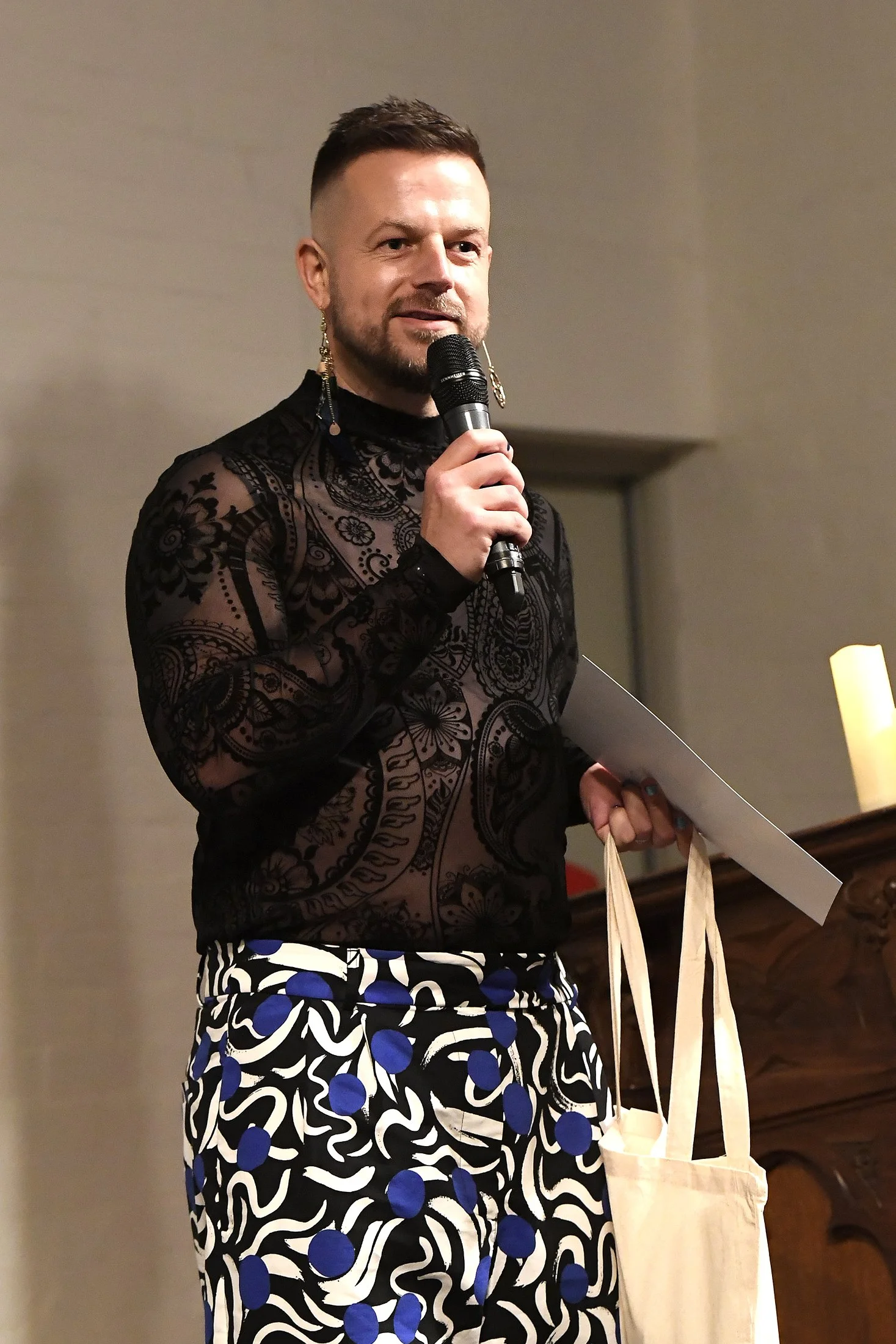 Person speaking into a microphone, wearing a see-through black lace top with intricate patterns and patterned pants with blue and white designs, holding a tote bag and a folder.