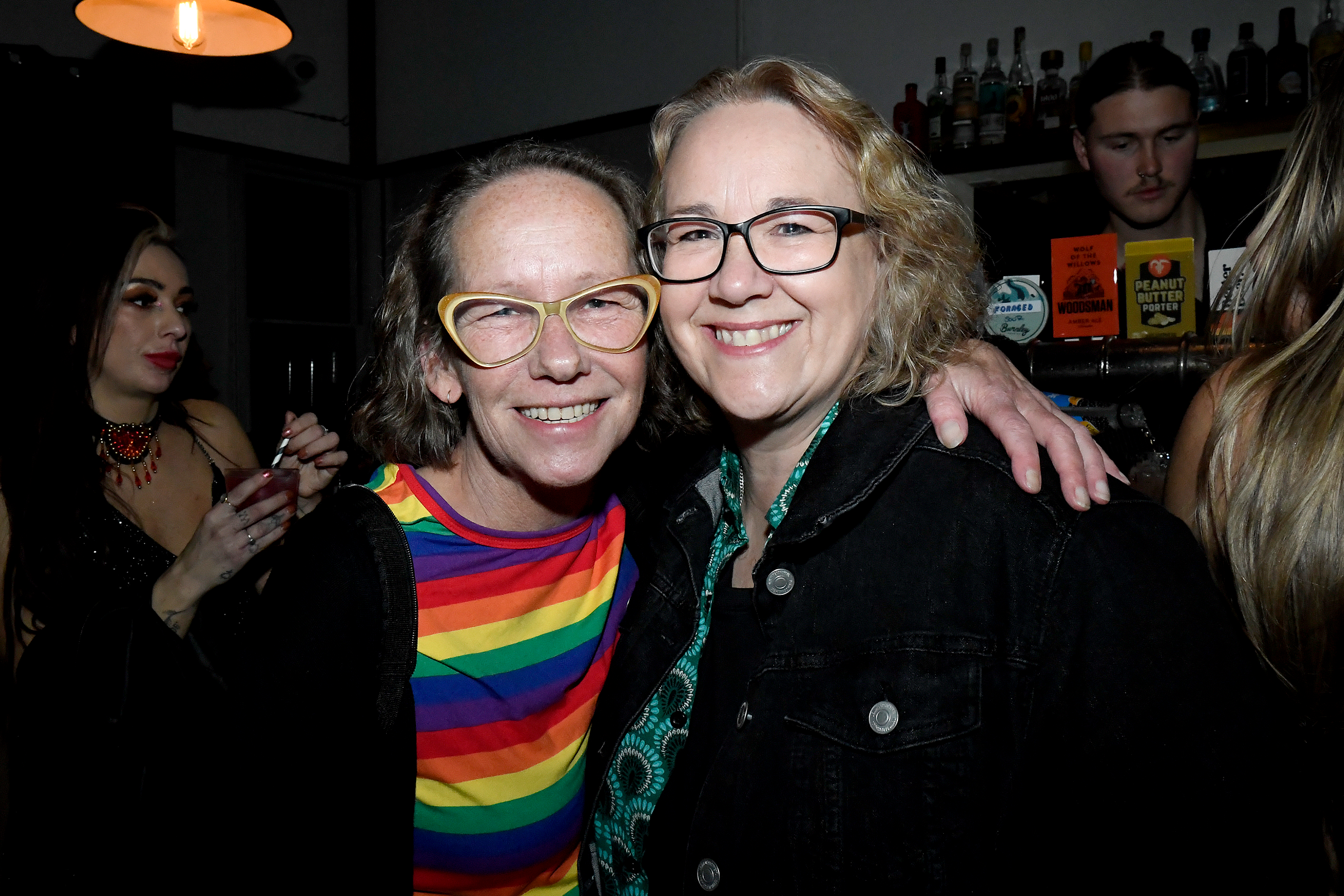 Two smiling women with glasses, one wearing a rainbow striped shirt and the other a black jacket, hugging each other at a social gathering. In the background, a woman with dark hair and makeup holds a drink, and a man with dark hair stands behind a b