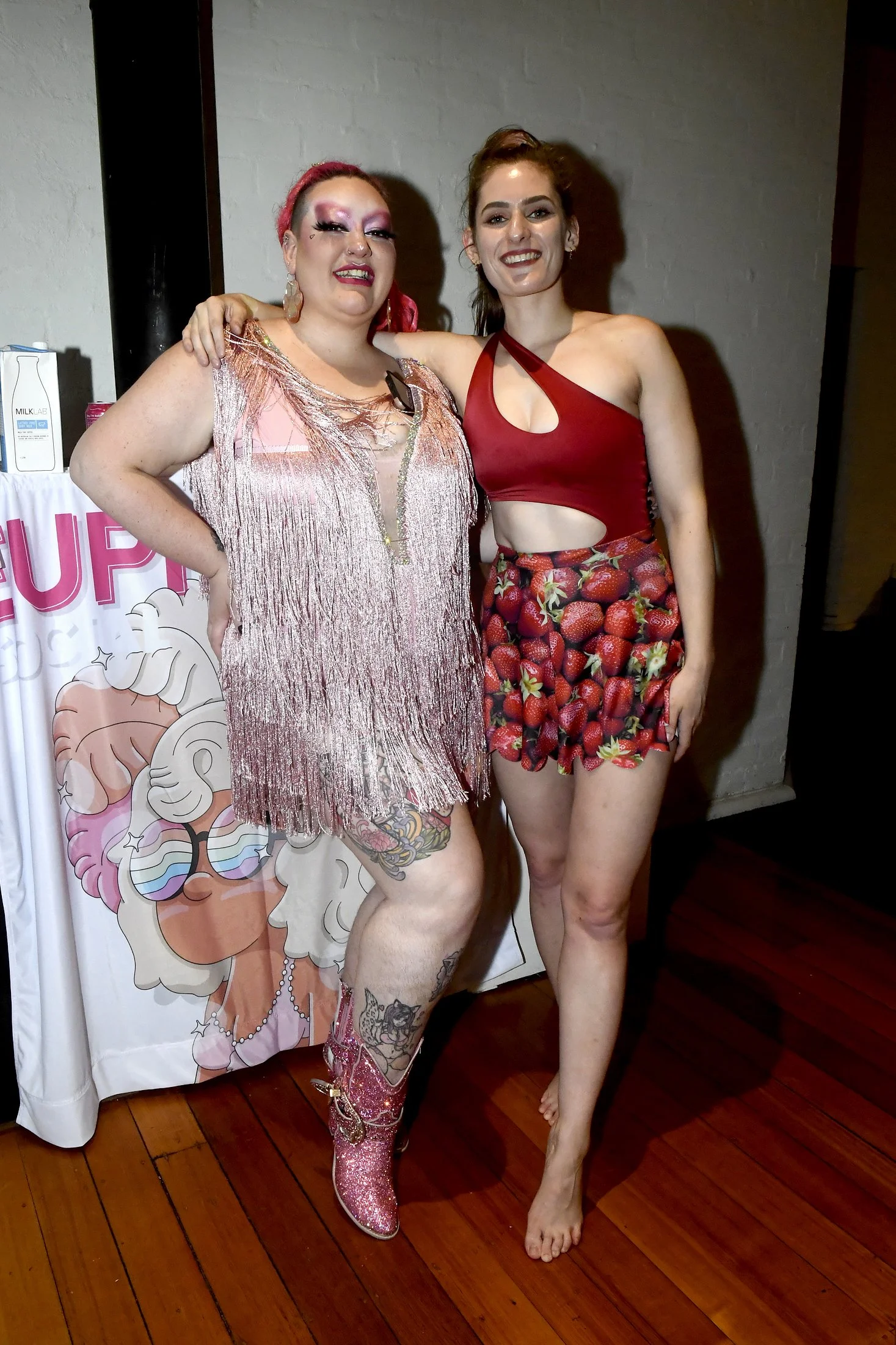 Two women standing together, one with rainbow-colored hair and tattoos wearing a shiny pink dress and cowboy boots, the other with brown hair wearing a red crop top and strawberry-patterned shorts. They are smiling and posing indoors.