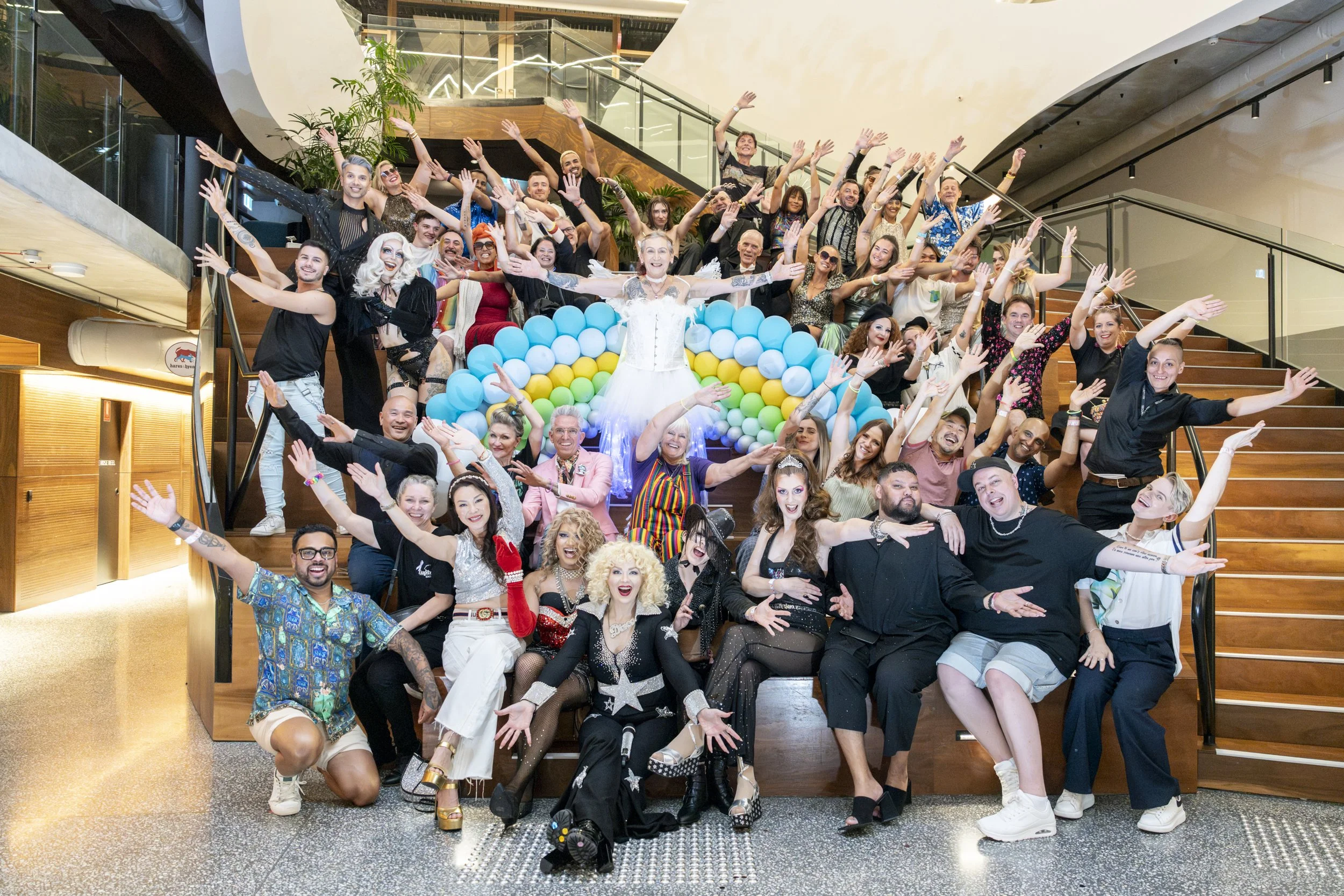 Large group of people celebrating on a staircase with a rainbow balloon arrangement, with a woman in a white dress at the center and everyone smiling and waving.