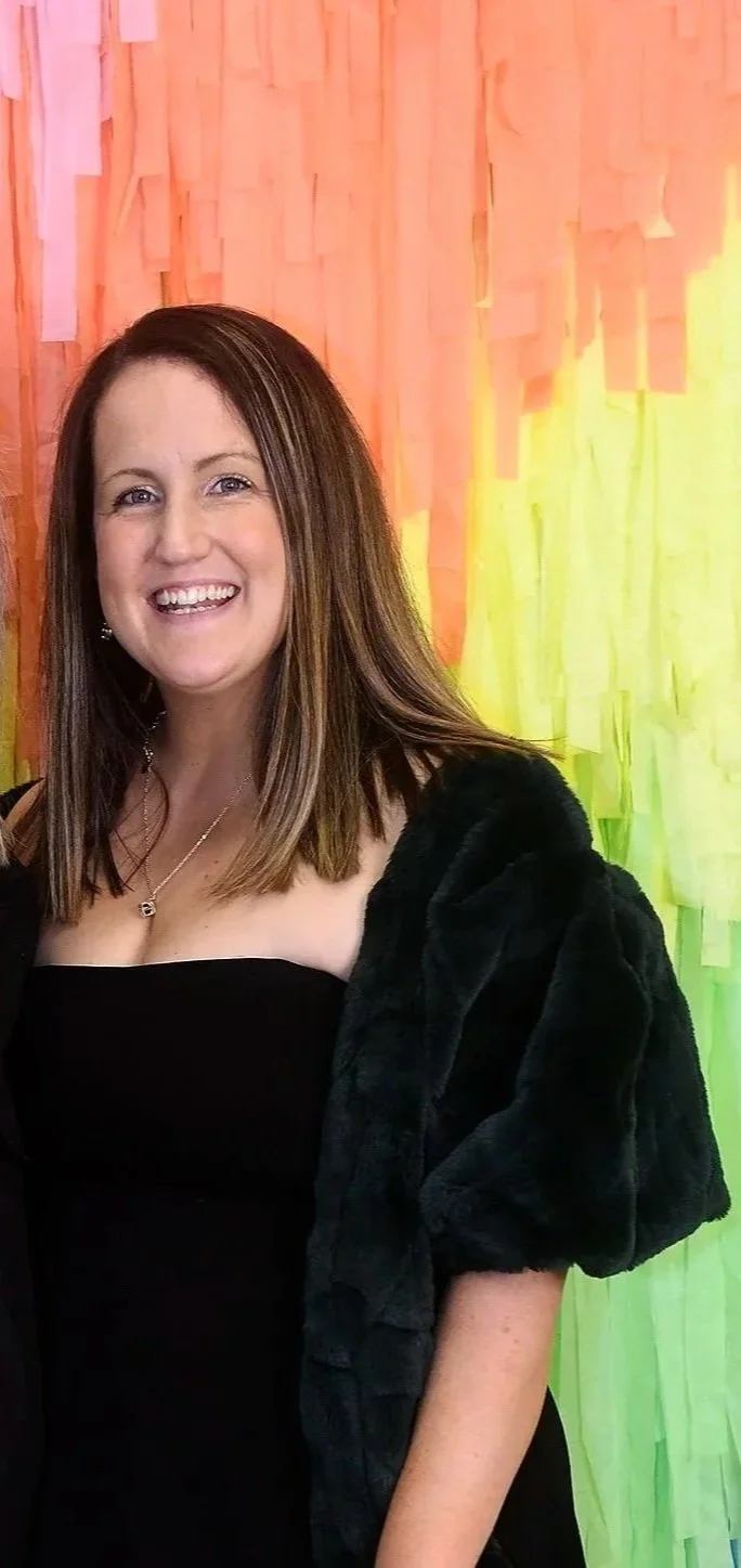 A woman smiling, wearing a black strapless dress and a black shawl, standing in front of a colorful backdrop with orange, yellow, and green paper flowers.