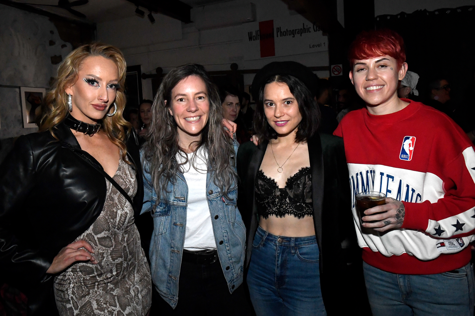 Four women standing together at an indoor event, smiling at the camera. The first woman on the left has long, wavy blonde hair and is wearing a black leather jacket over a patterned dress. The second woman has long, curly gray hair and is dressed in 