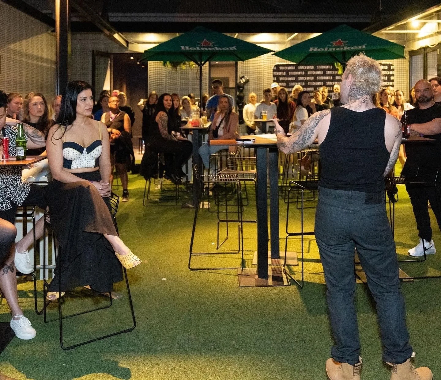 A man with tattoos on his arms and neck is speaking to a woman seated in a black and white outfit in a crowded outdoor bar or restaurant at night. The crowd is sitting and standing, and there are large green umbrellas with Heineken logos overhead.