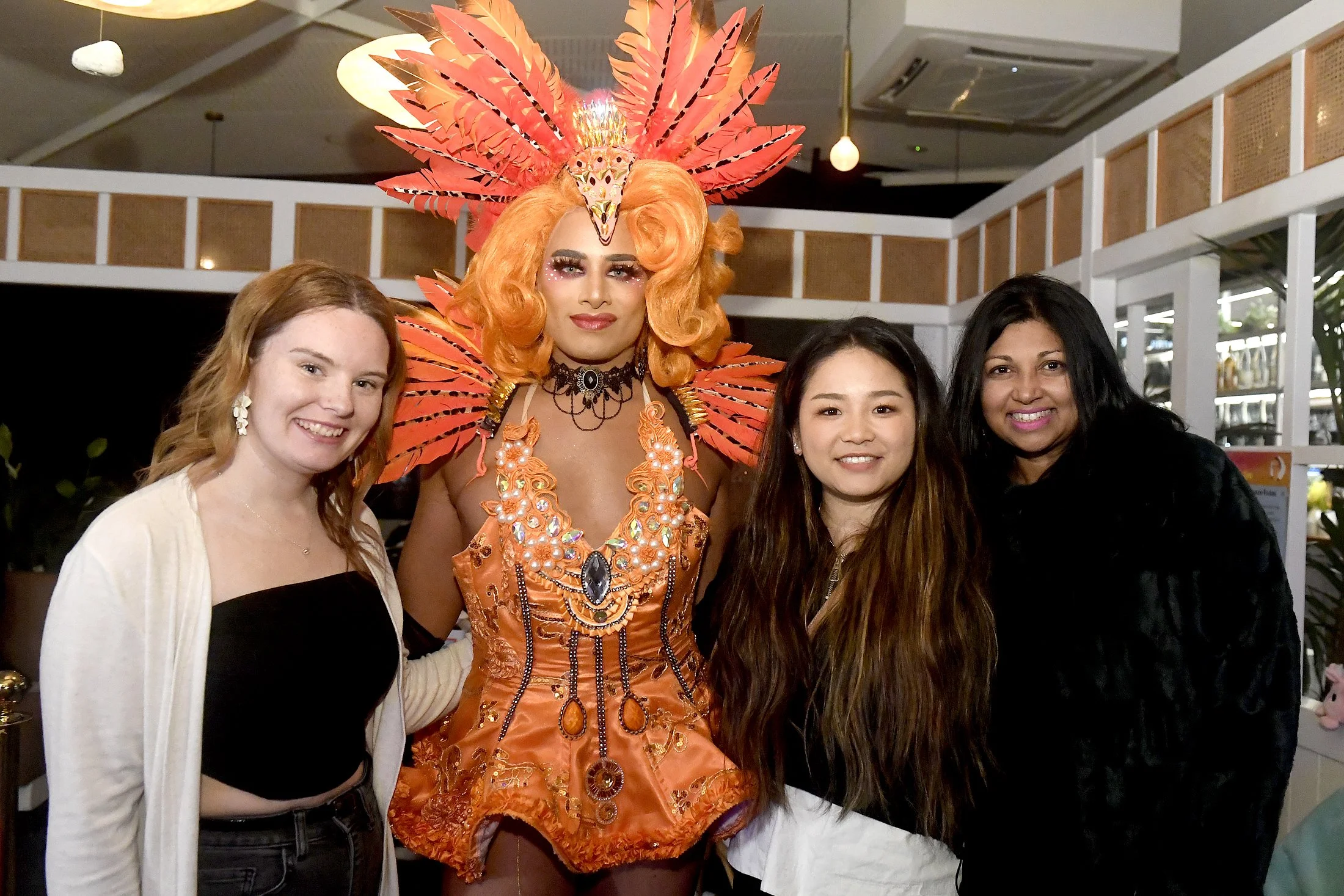 A group of four women and a drag queen dressed in an elaborate orange costume with feathers and jewelry, posing together indoors.