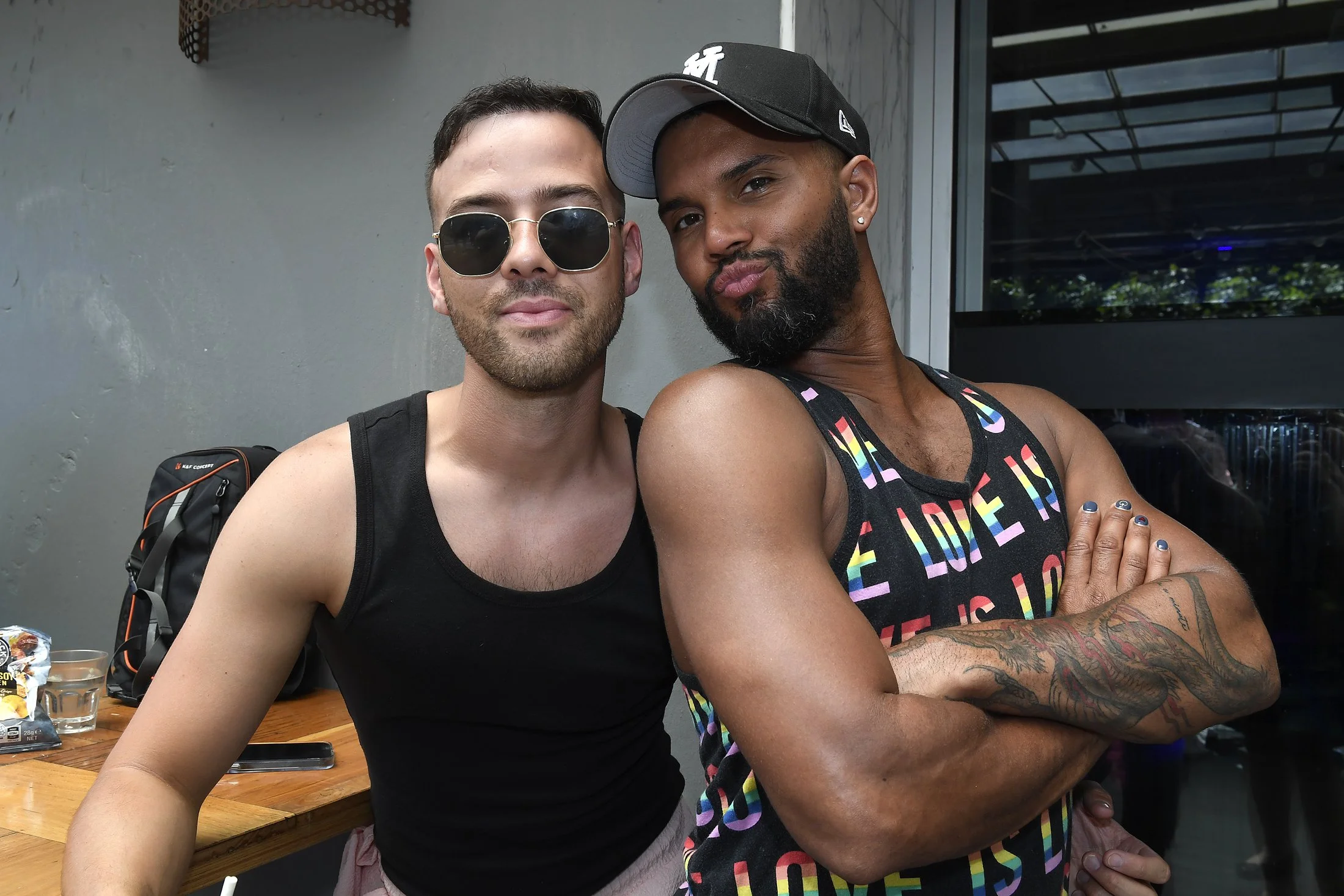 Two men posing together indoors; the man on the left wears sunglasses and a black tank top, the man on the right has a beard, a black cap, colorful tank top, and tattoos on his arm, standing with arms crossed.