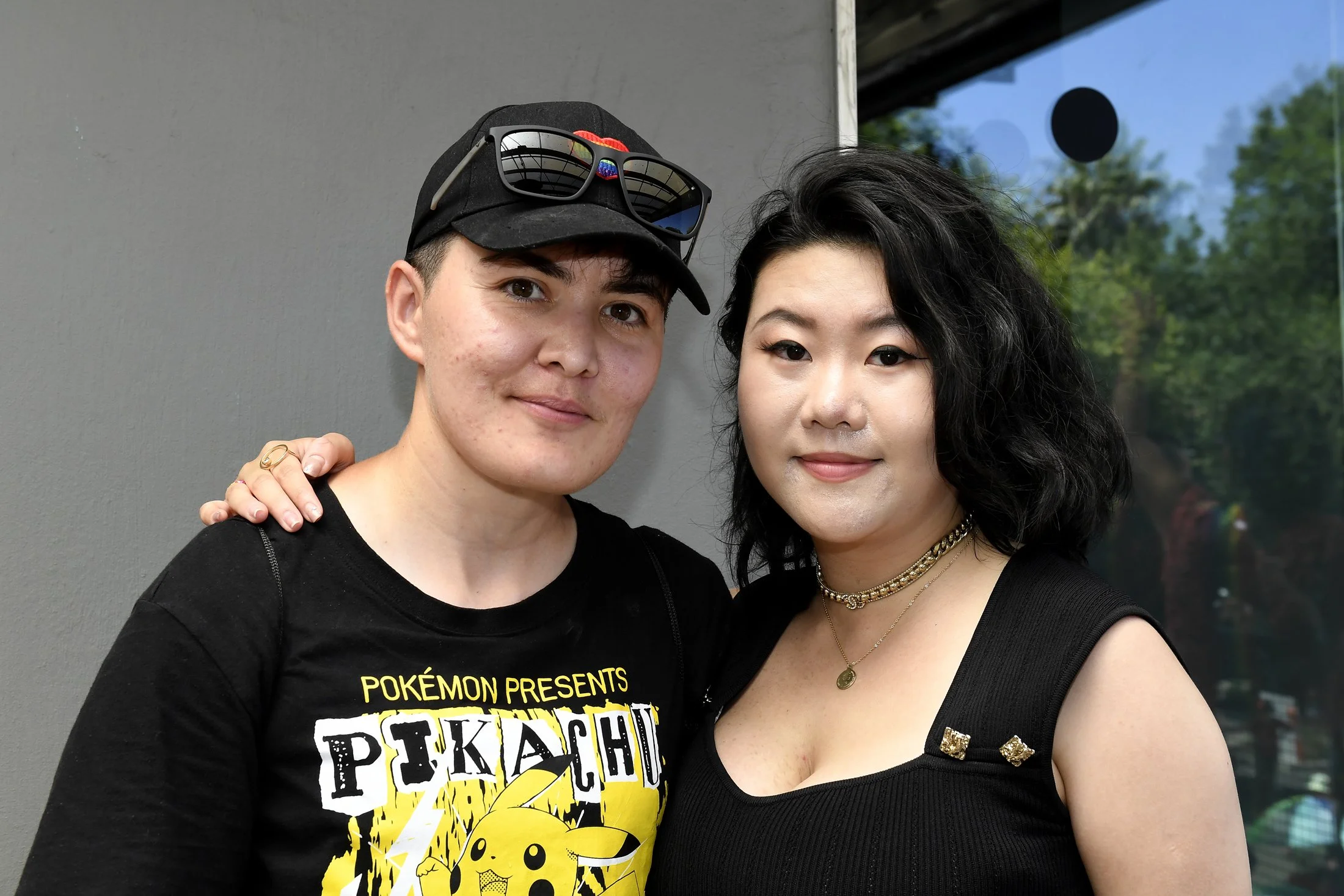 Two women standing closely together, with the woman on the left wearing a Pikachu t-shirt, black cap with sunglasses, and the woman on the right wearing a black top and layered necklaces, both smiling at the camera.