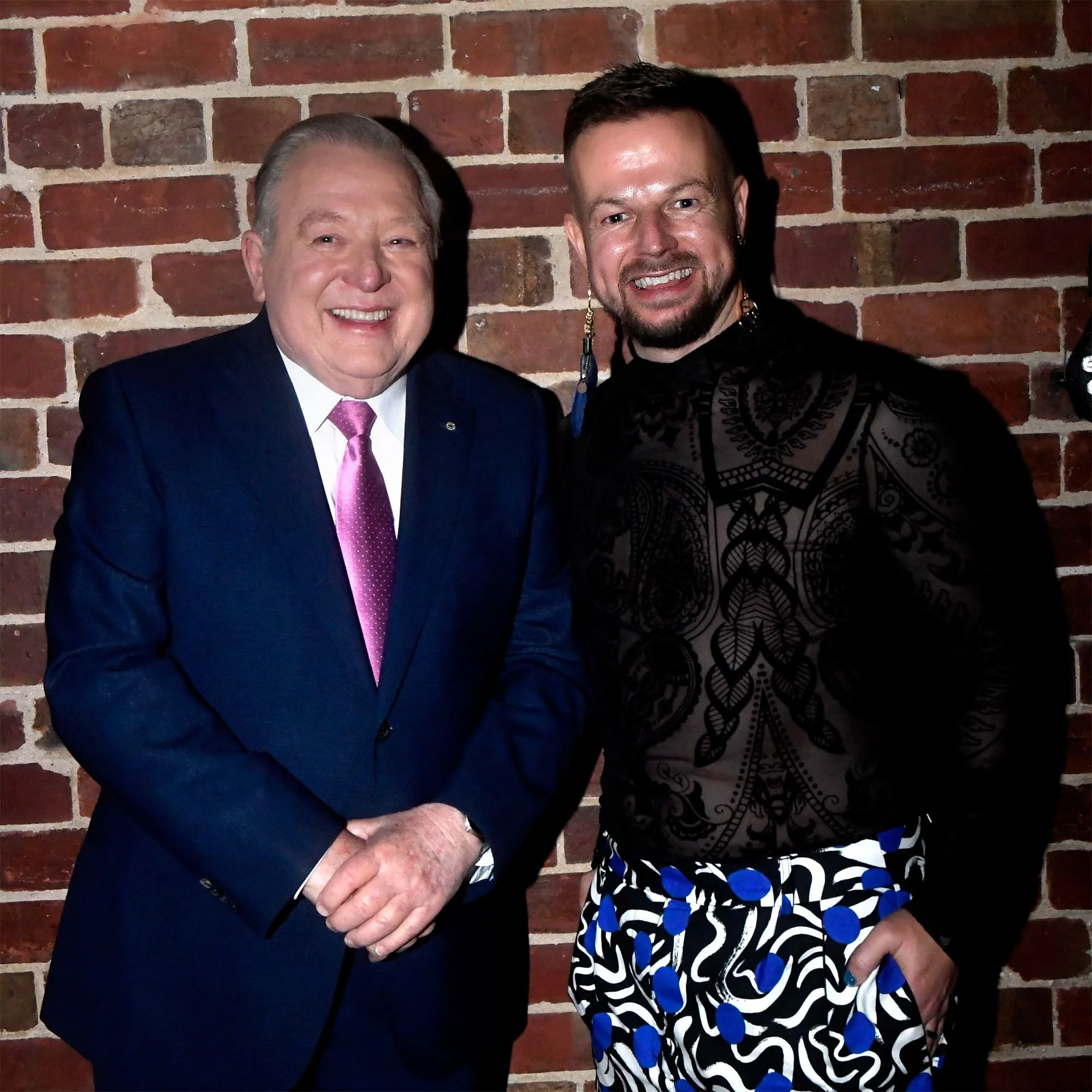 Two men standing in front of a brick wall, smiling. The man on the left is wearing a navy suit with a pink tie, and the man on the right is wearing a black shirt with patterned black and blue pants.