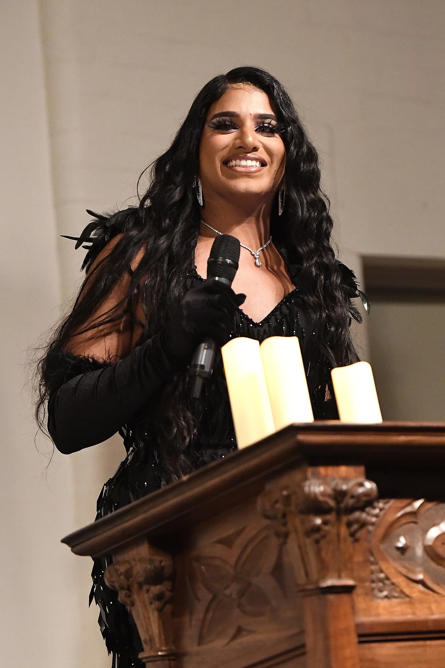 A woman with long black curly hair wearing a black dress and gloves, holding a microphone, standing at a wooden podium with candles.