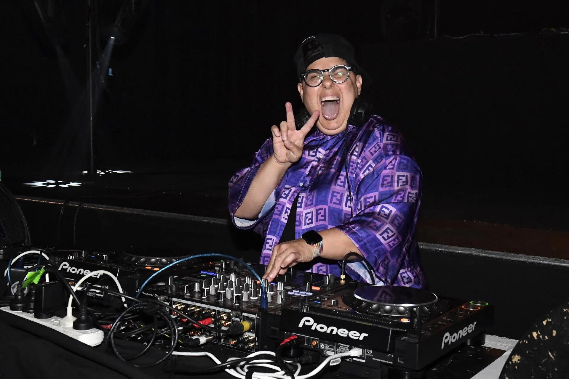 Person DJing at a club, wearing a purple patterned shirt, black hat, glasses, making a peace sign, open mouth, standing behind DJ equipment.