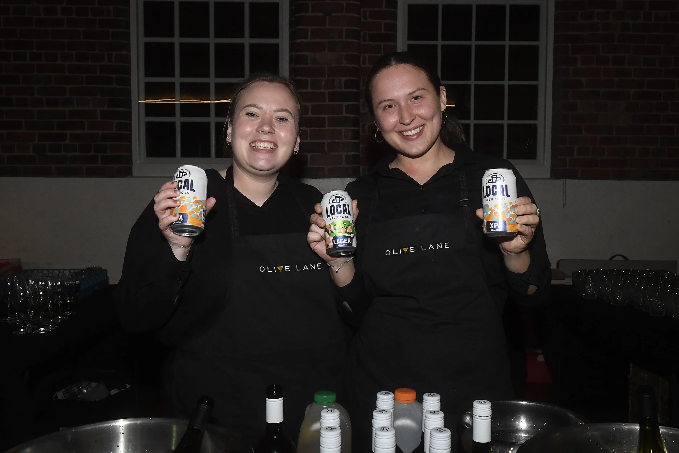 Two women smiling and holding cans of beer at a bar. They are wearing black aprons with 'Olive Lane' written on them. The background shows a brick wall and a window.