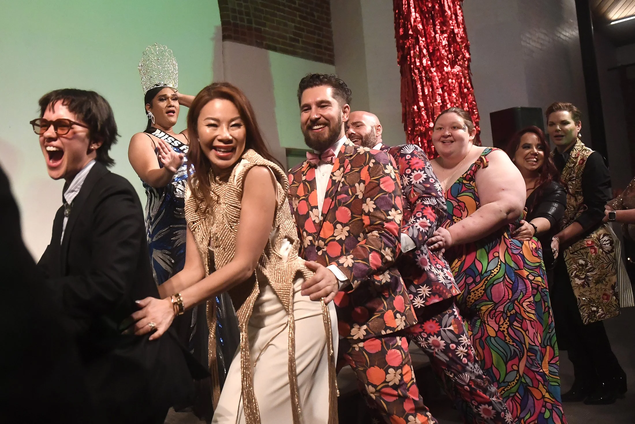 A diverse group of people participating in a conga line at a lively event with colorful attire, including floral suits and patterned dresses, under a red sequin curtain.