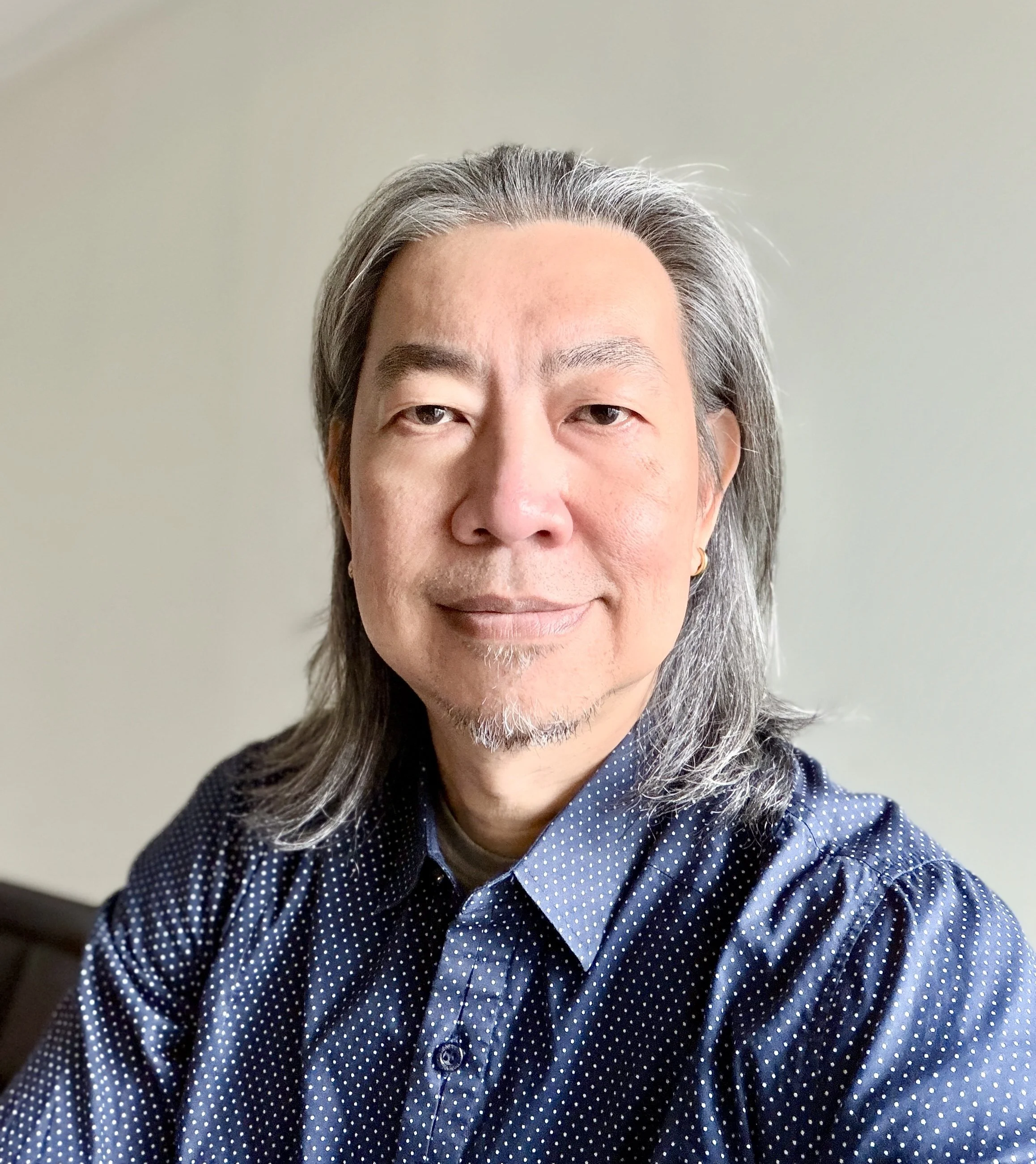 Close-up of a middle-aged Asian man with salt-and-pepper hair, wearing a navy blue polka-dot shirt, smiling softly, indoors with plain light-colored wall background.