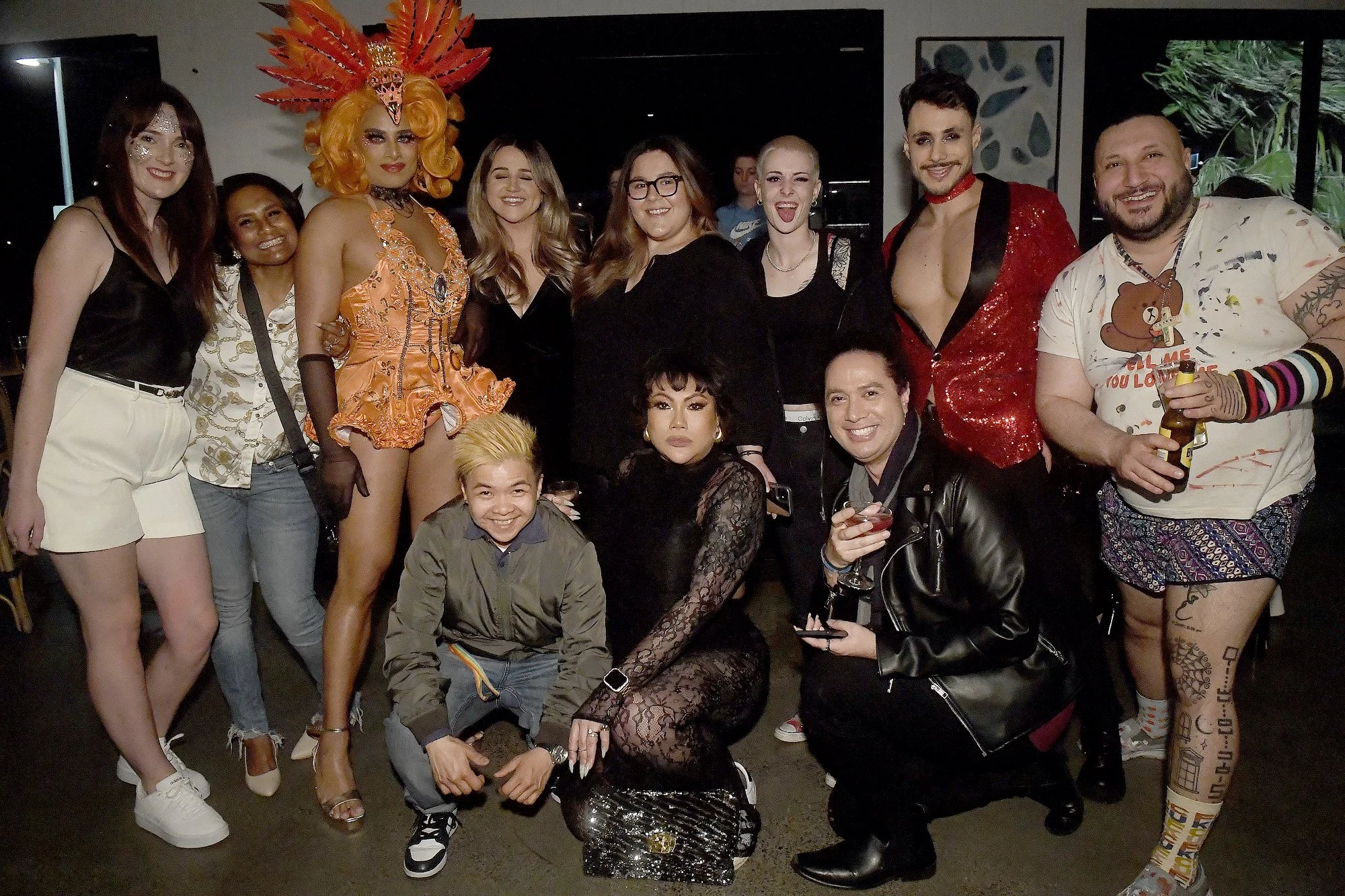 Group of diverse friends at a celebration, some in colorful costumes, posing indoors with drinks, smiling and showing playful expressions.