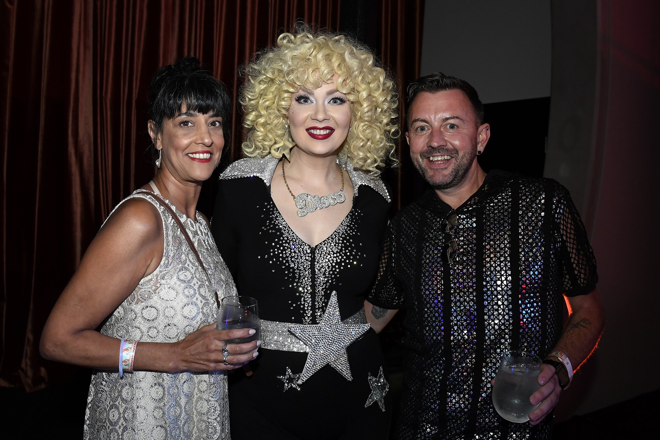 Three people smiling and posing for a photo at a party. The woman on the left wears a silver dress and holds a glass. The person in the middle has curly blonde hair, a black outfit with silver stars, and a large star belt buckle. The man on the right