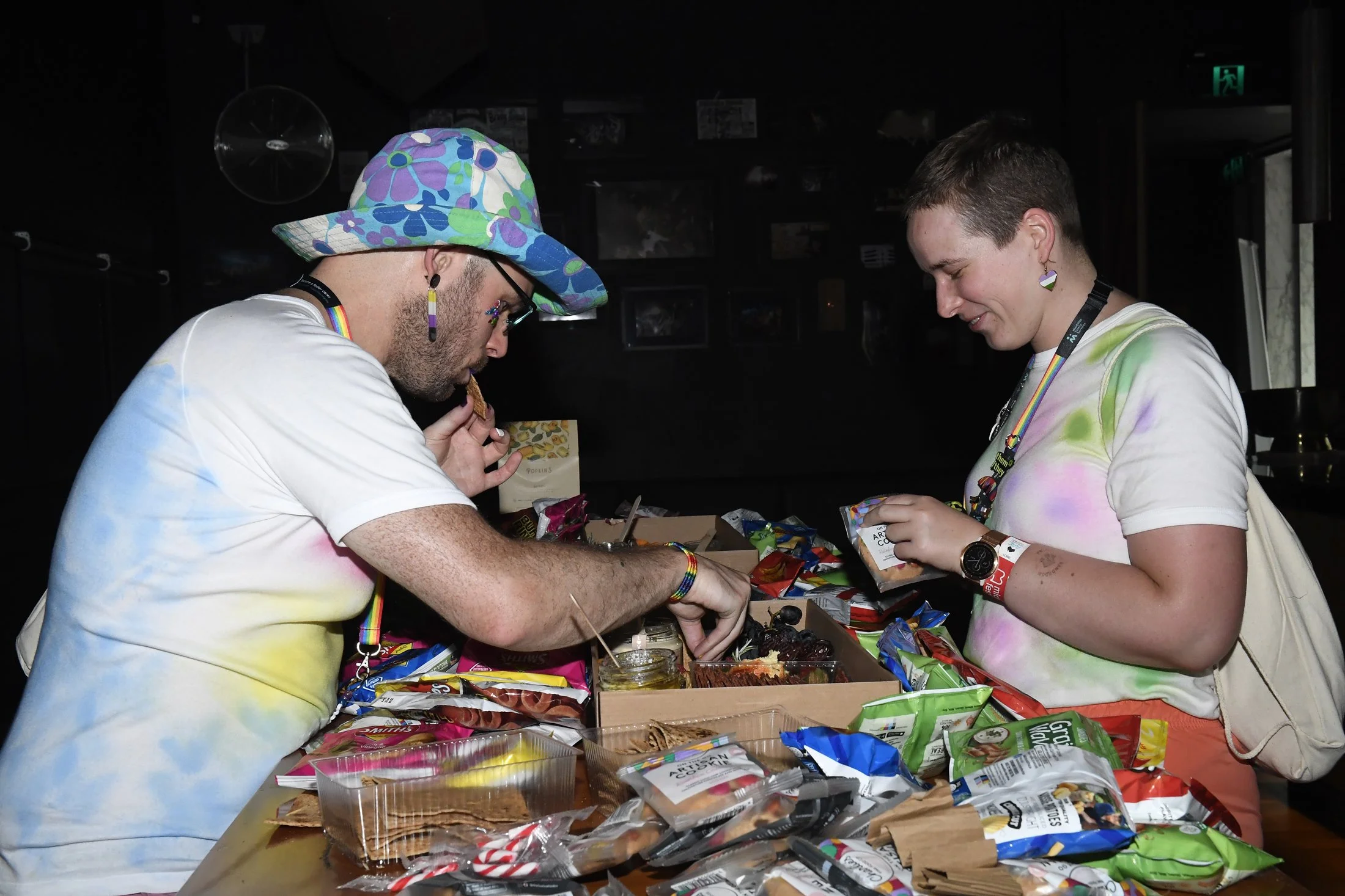 Two people at a table filled with snacks, both wearing rainbow-colored accessories and colorful tie-dye shirts, browsing and selecting snacks.