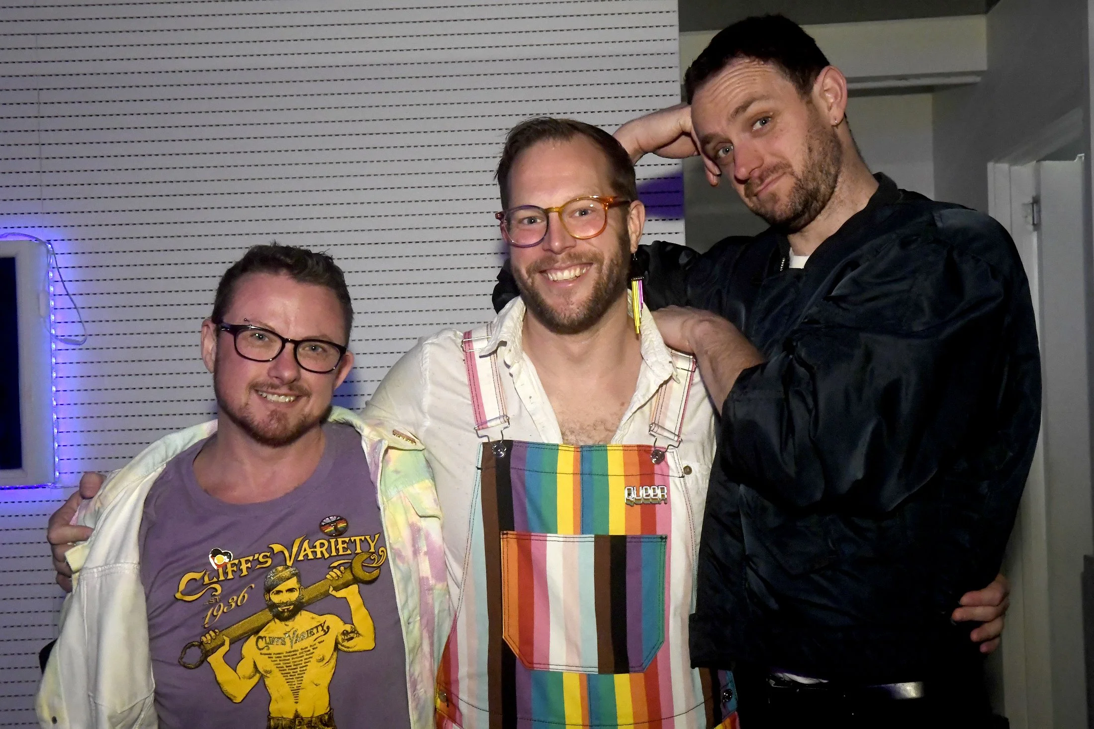 Three men smiling and posing together indoors. The man on the left wears glasses and a graphic T-shirt. The man in the middle has glasses, a beard, and wears a colorful rainbow apron. The man on the right has dark hair and a beard, wearing a black ja