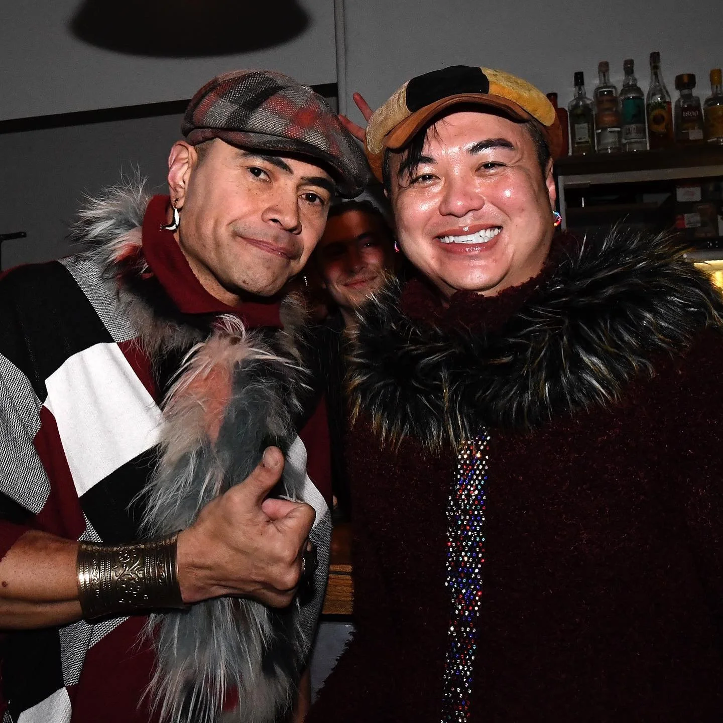 Two men smiling at the camera, one giving a thumbs-up, indoors with shelves of bottles in the background.