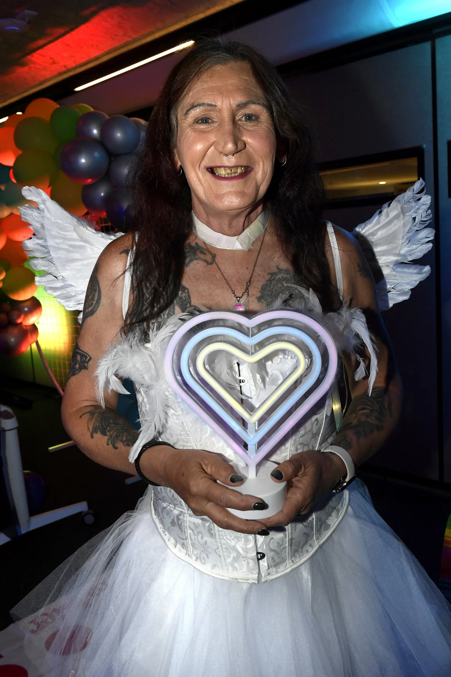 A smiling woman with dark hair, tattoos, and a septum piercing, dressed in a white dress with angel wings, holding a heart-shaped neon light at a colorful party or celebration.