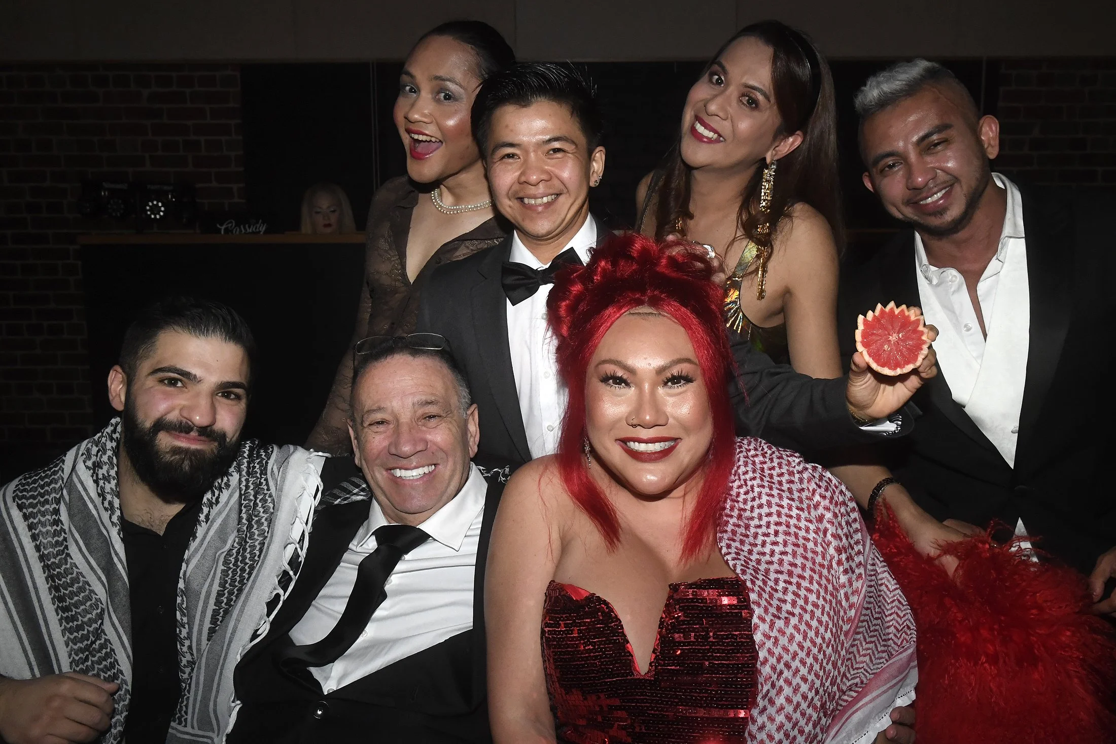 Group of nine diverse adults dressed at a party, smiling, with a woman holding a grapefruit slice and wearing bright red hair.