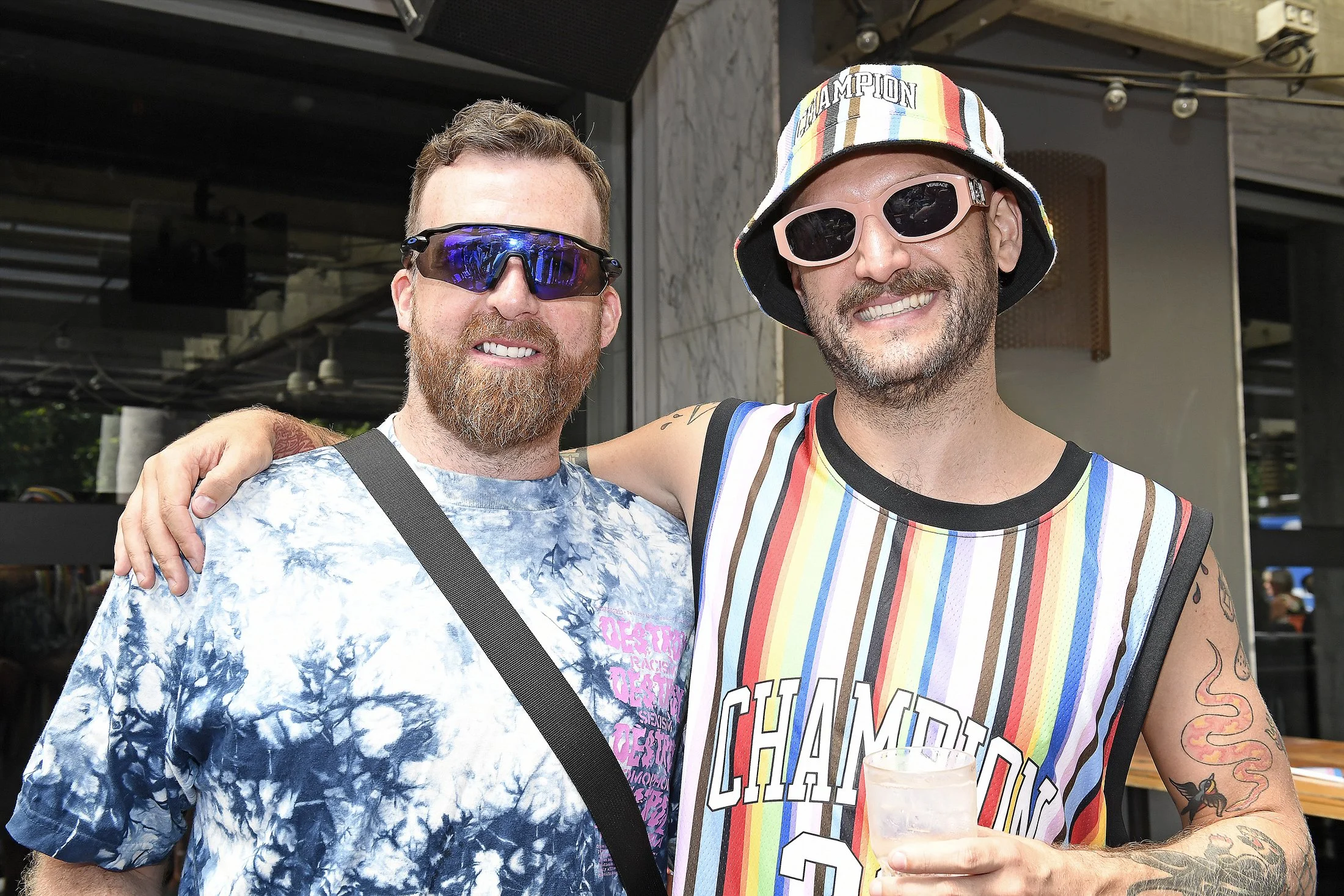 Two men smiling, one with a beard and sunglasses, wearing a blue tie-dye shirt, and the other with a beard, wearing a colorful bucket hat, pink sunglasses, and a rainbow-striped basketball jersey holding a drink, standing together outdoors.