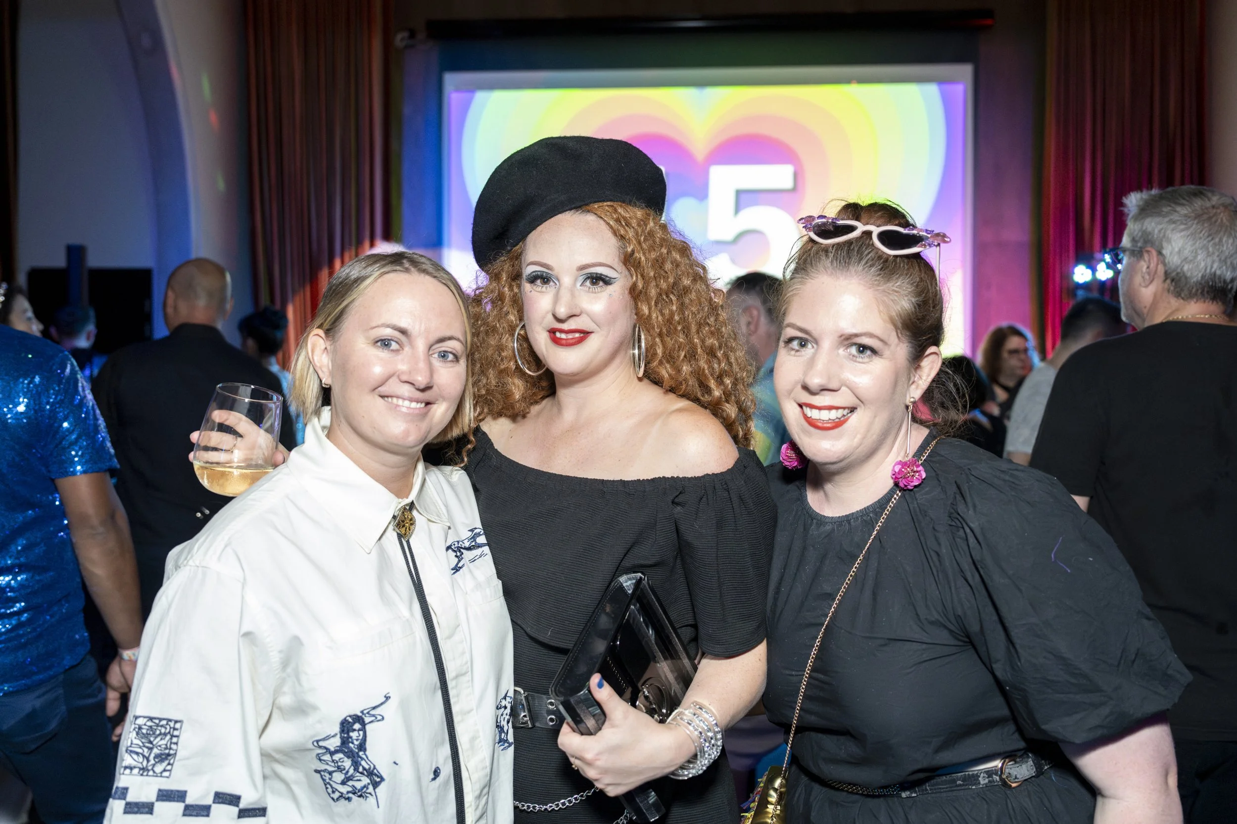 Three women smiling at a party, standing close together. The woman on the left has blonde hair, light skin, and is holding a wine glass. The woman in the middle has curly red hair, fair skin, and is wearing a black off-shoulder dress and a black bere