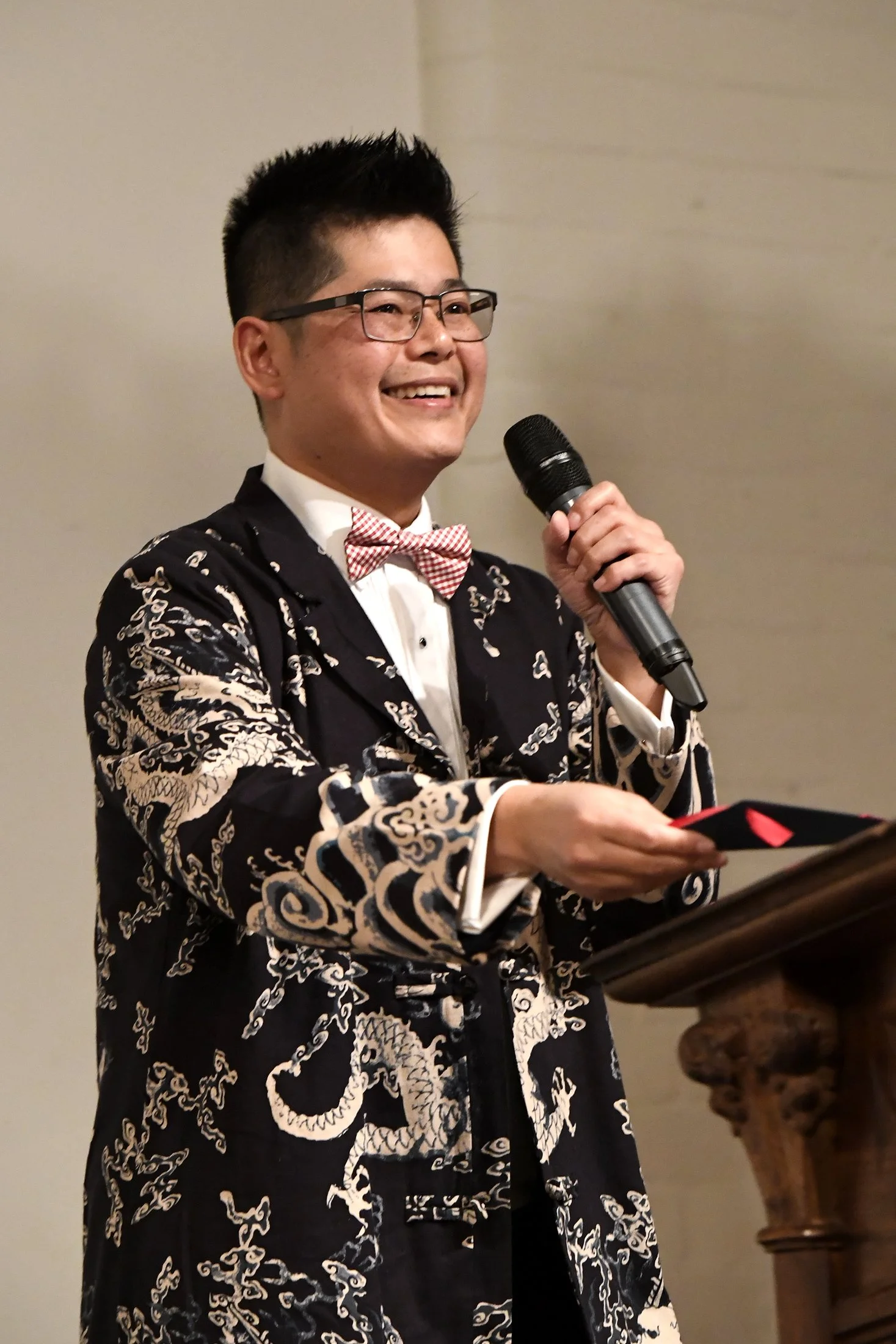 A man wearing glasses, a red and white checkered bow tie, and a black robe with intricate white and gold dragon designs, standing at a podium holding a microphone and a small black card, smiling during a speech or presentation.