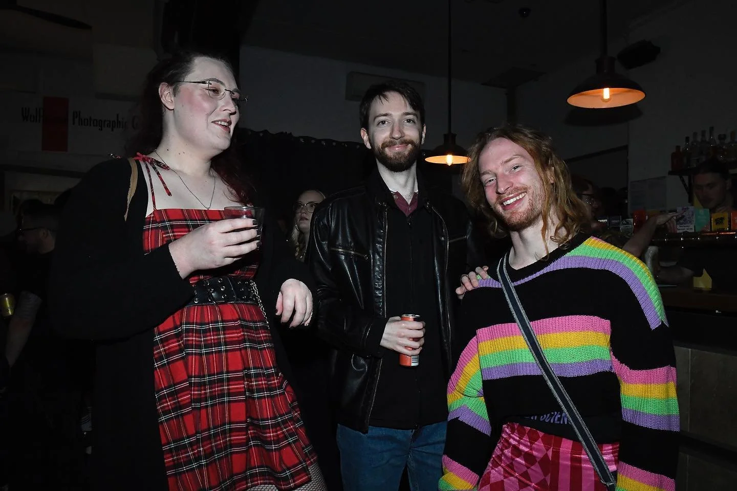 Three friends at a bar, two men and one woman, smiling and holding drinks. The woman wears glasses and a red plaid dress. One man has a beard and wears a black leather jacket. The other man has long red hair, a beard, and wears a colorful striped swe