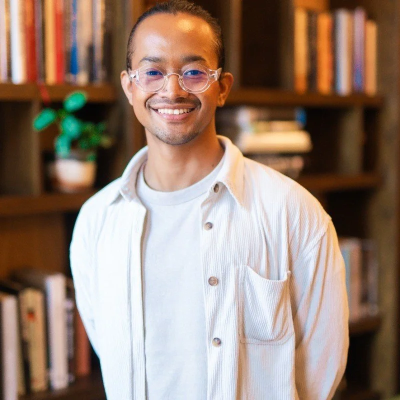 A young man with glasses and a smile standing in front of a bookshelf filled with books and a potted plant.