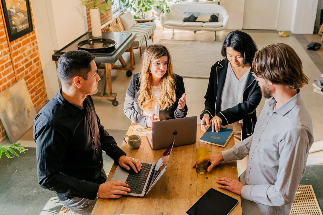 Four employees stand at a conference table with their laptops and have a discussion.
