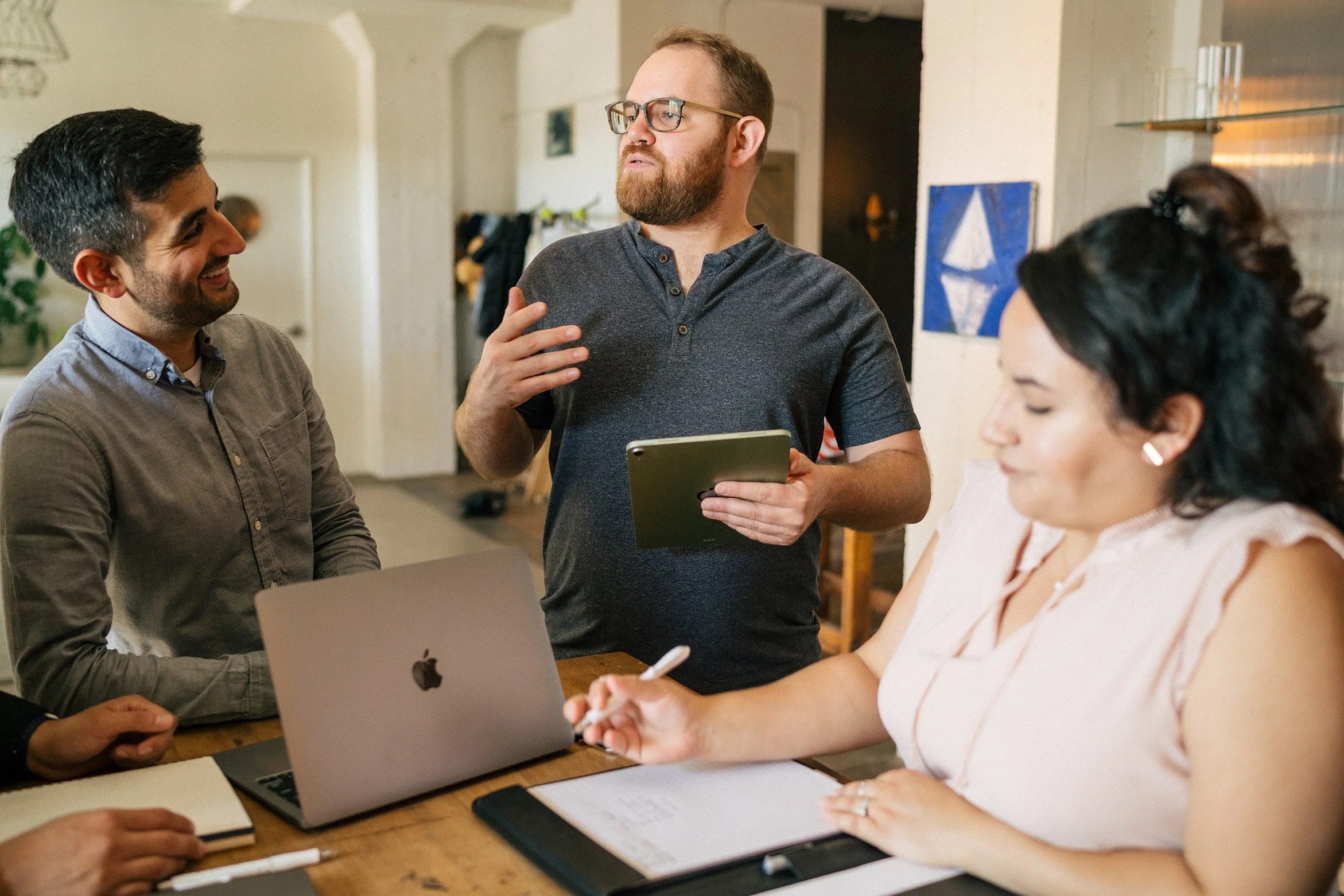 Man Three employees stand around a conference table.  A man holds an ipad and presents information as teammates listen and take notes.