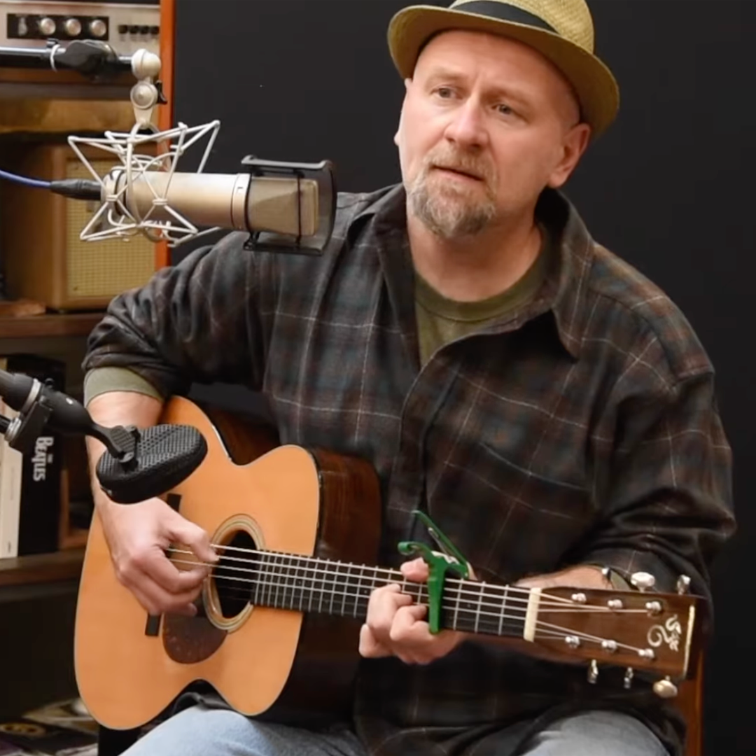 A man wearing a plaid shirt and a straw hat is playing an acoustic guitar and singing into a microphone in a recording studio.