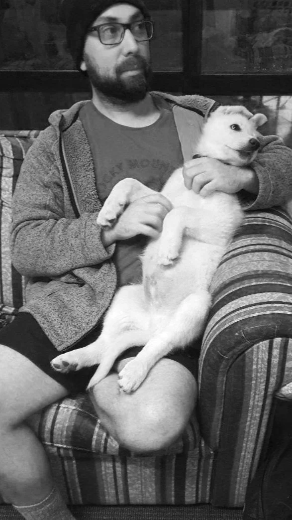 A man with glasses and a beard sitting on a striped armchair, holding a puppy in his lap. The man is wearing a beanie, glasses, a t-shirt, and a zip-up hoodie. The puppy appears to be a light-colored breed, lying on its back with its head resting on the man's arm.