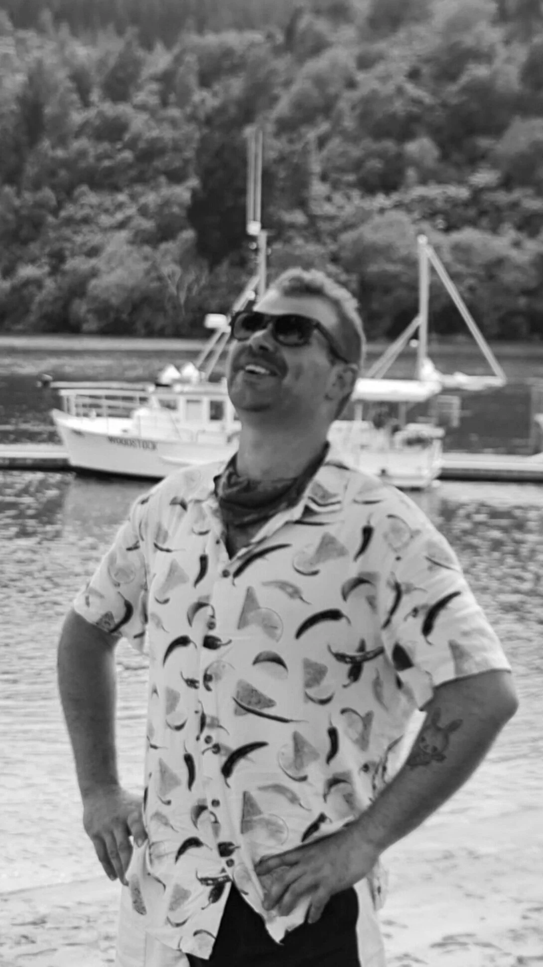 A man stands on a beach with his hands on his hips, wearing sunglasses, a patterned shirt, and a bandana around his neck. There is a river or lake with boats and a wooded hillside in the background.